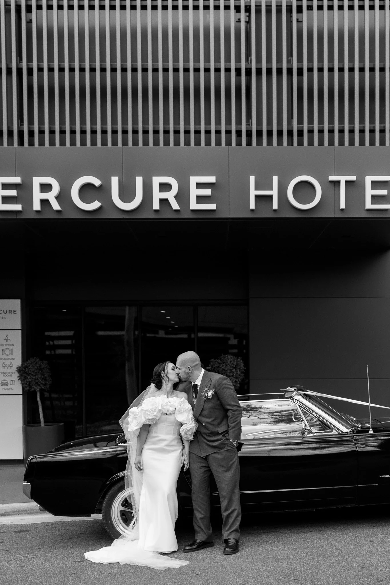 A bride and groom kissing in front of a classic black car outside the Mercure Hotel.