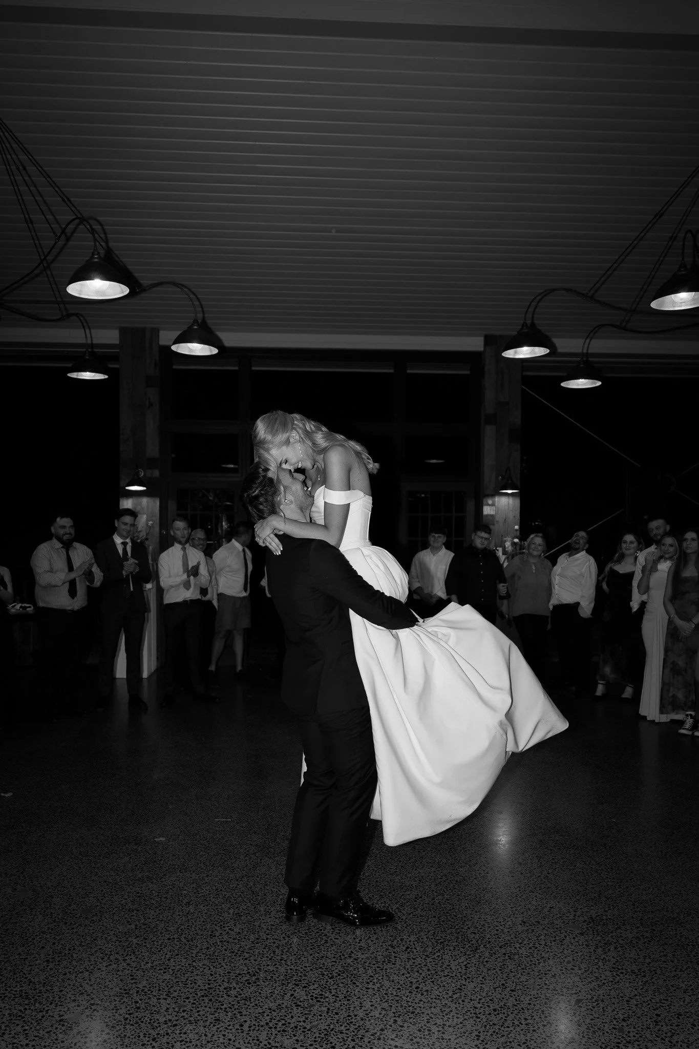 A black-and-white photo of a wedding reception where a groom is lifting the bride in the air. The bride is wearing a white wedding gown and the groom is dressed in a black suit. Guests in formal attire are watching and applauding in the background in