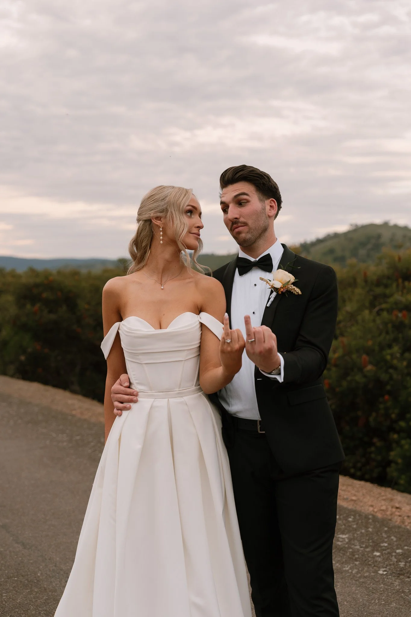 A bride and groom stand together outdoors, with the bride in a white wedding dress and the groom in a black tuxedo, both showing their middle fingers to the camera.