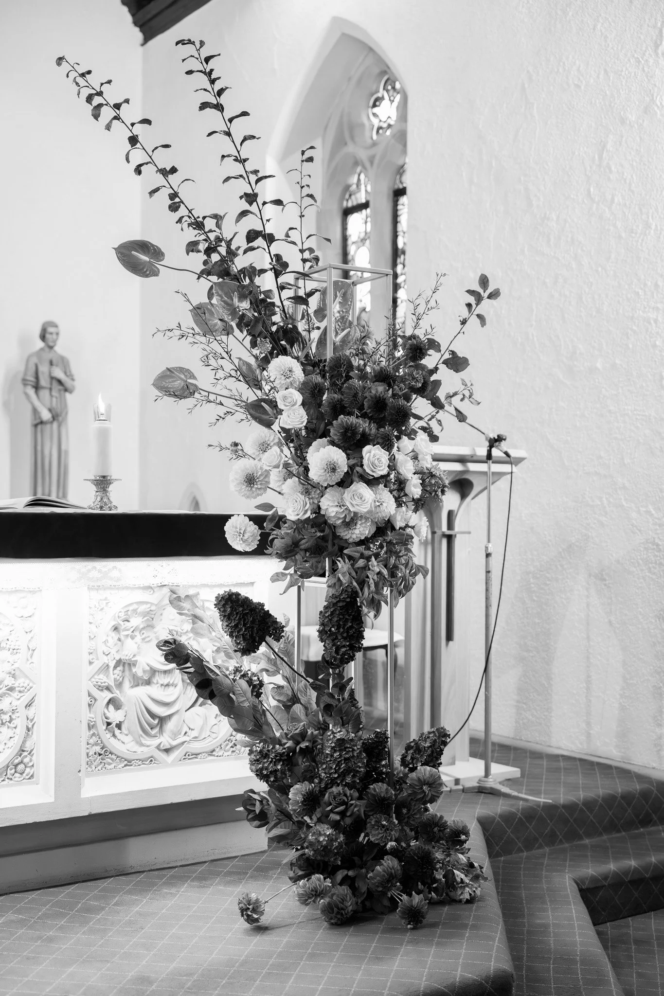Black and white photo of a floral arrangement on a church altar, with statues and stained glass windows in the background.
