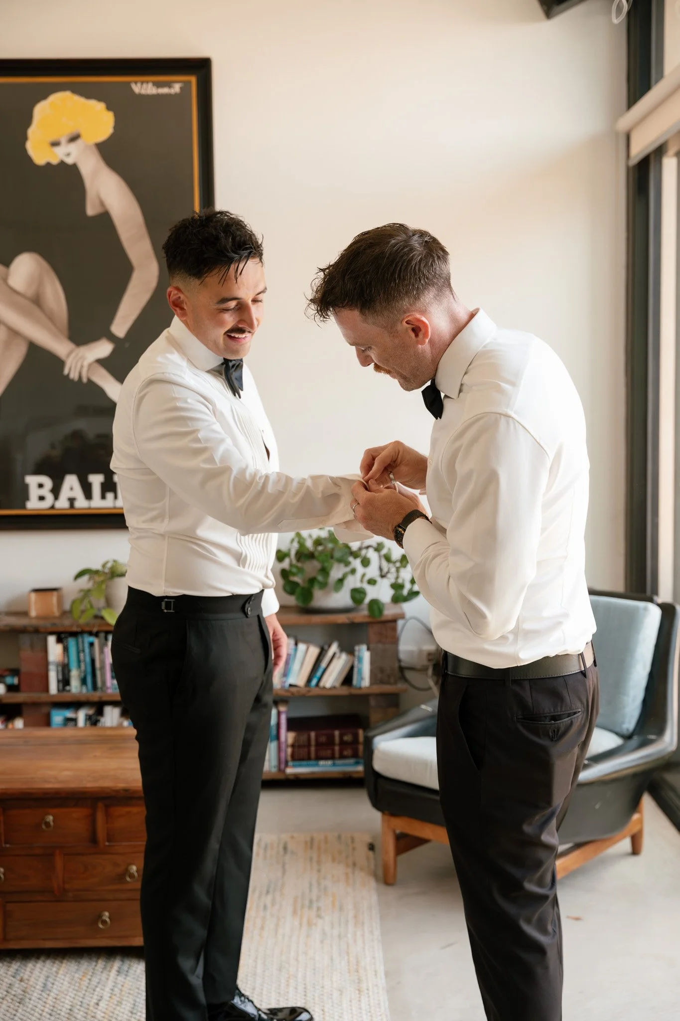 Two men dressed in white shirts and black pants in a living room, one is helping the other with cufflinks, by a window with natural light and a poster on the wall.