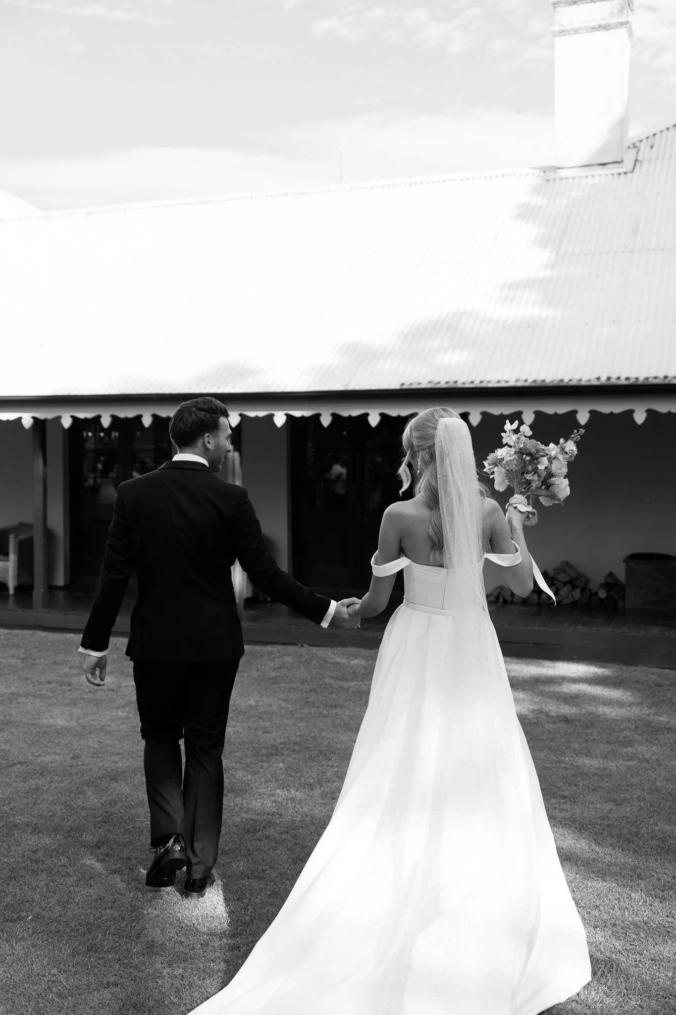 A bride and groom walk hand in hand on a grassy area outside a building, with the bride holding a bouquet of flowers, captured in black and white.