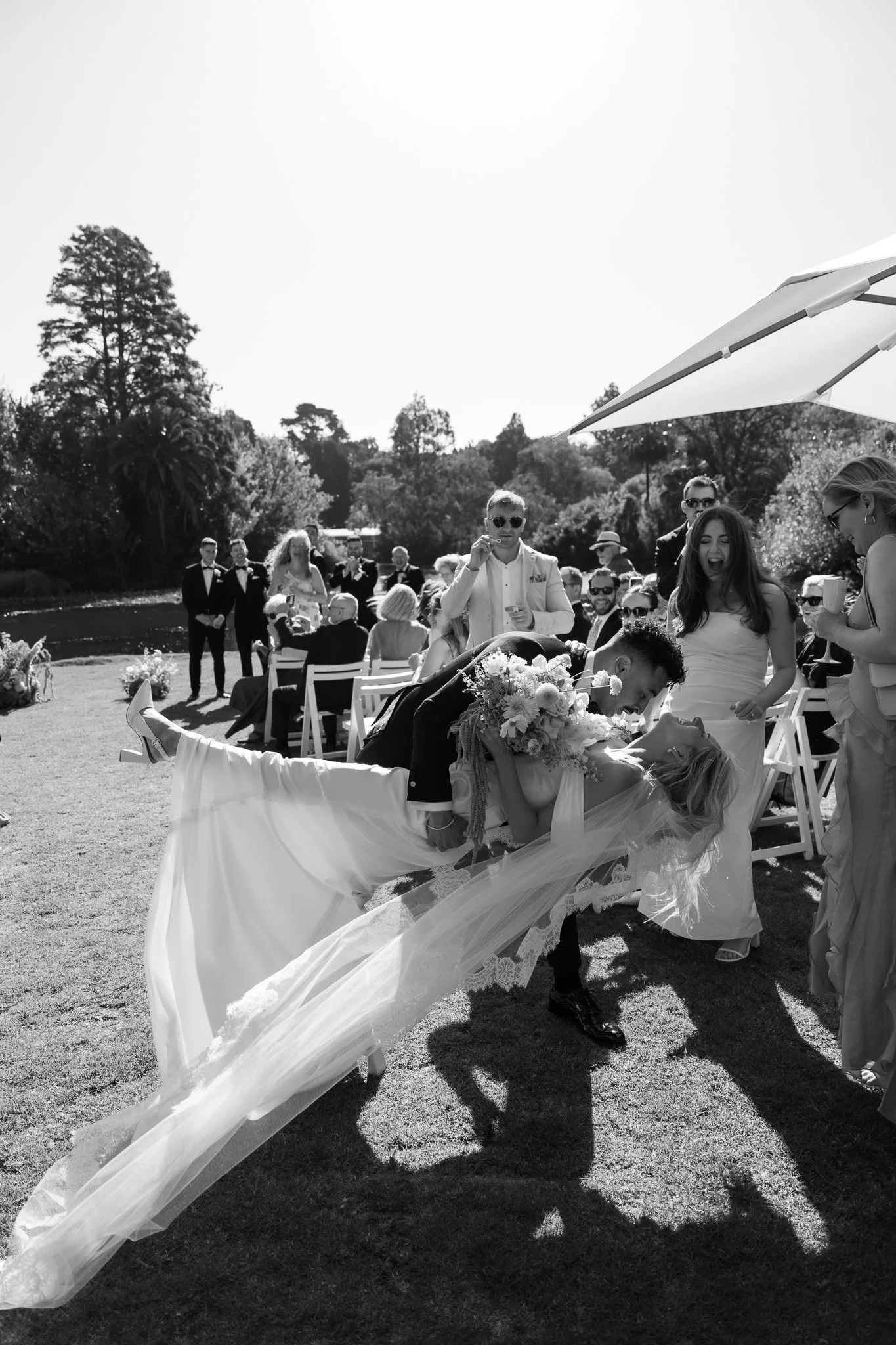 Bride and groom dancing at an outdoor wedding with guests watching and smiling, trees and a body of water in the background, black and white photo.