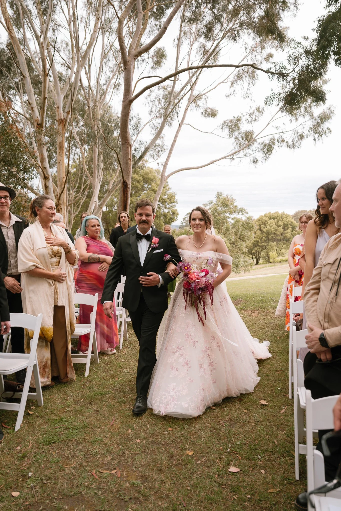 Bride and groom walking down the aisle at an outdoor wedding ceremony. The bride wears a strapless, off-the-shoulder wedding gown and holds a colorful bouquet of flowers. The groom wears a black tuxedo with a bow tie and boutonniere. Friends and fami