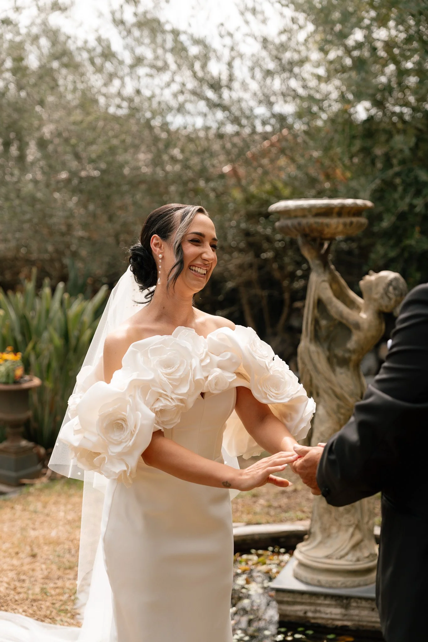 A bride in a white wedding dress with large floral sleeves, smiling and holding hands with a groom during a wedding ceremony outdoors.