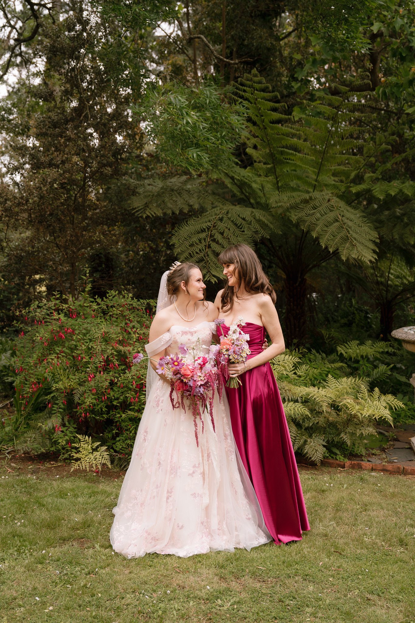 Two women in wedding and bridesmaid dresses holding bouquets, smiling and looking at each other outdoors in a garden setting.
