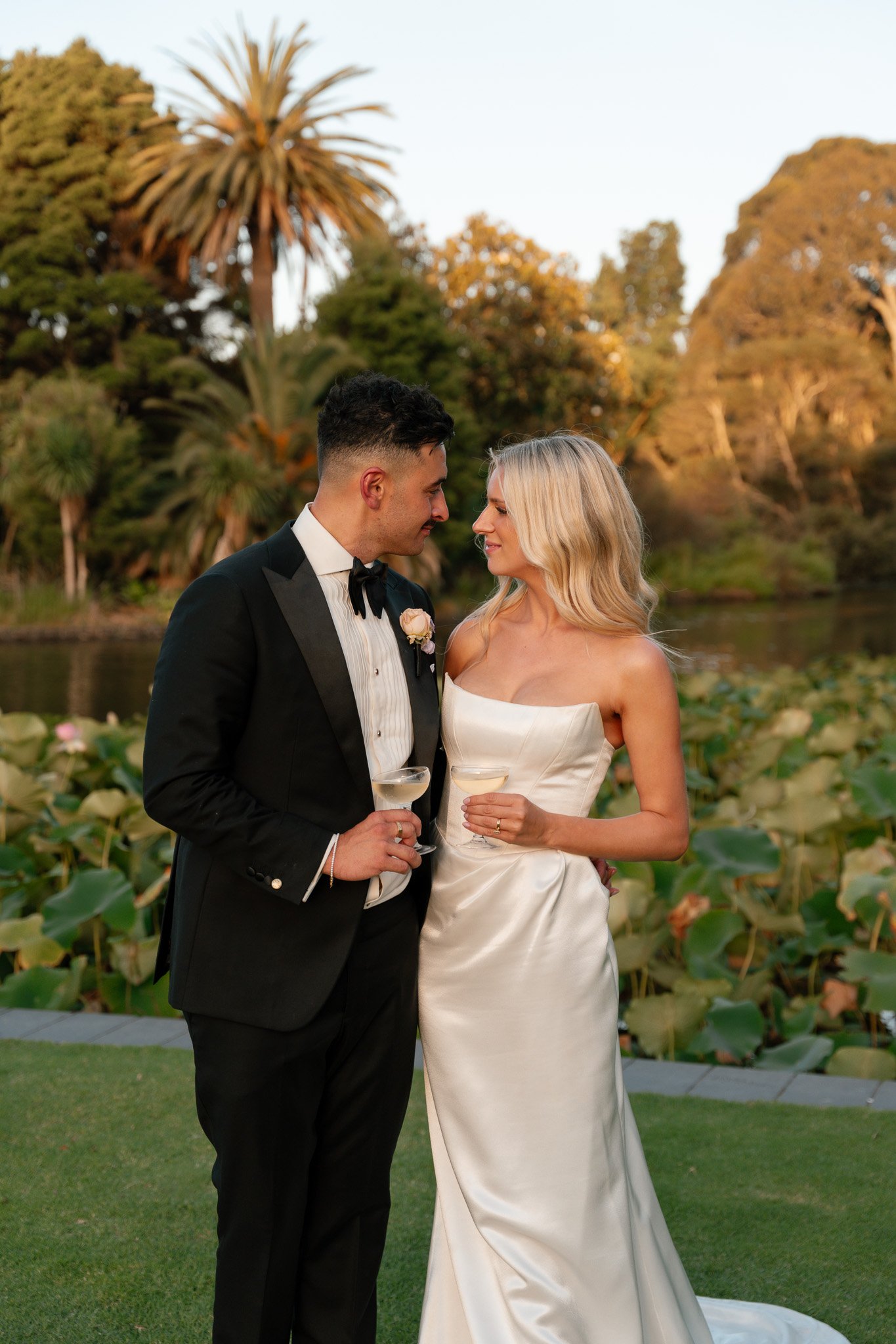 A newlywed couple, a man in a tuxedo and a woman in a white wedding dress, holding drinks and standing close together outdoors during sunset, with lush greenery and a pond in the background.