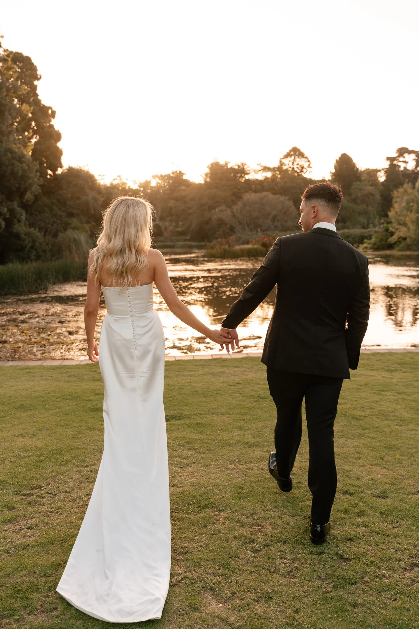 A bride and groom holding hands and walking near a lake during sunset.