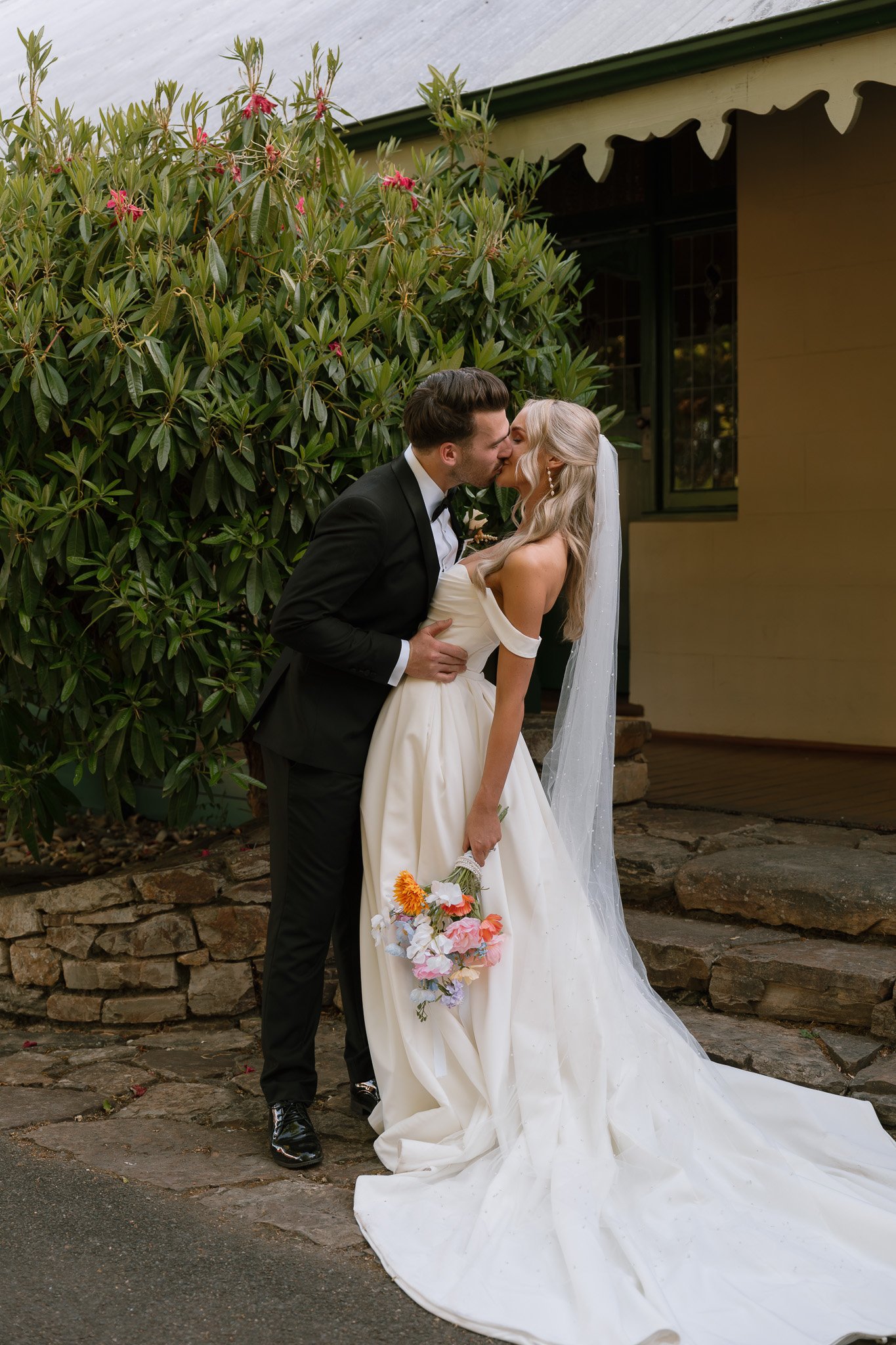 Bride and groom sharing a kiss outside, with the bride holding a bouquet of colorful flowers.