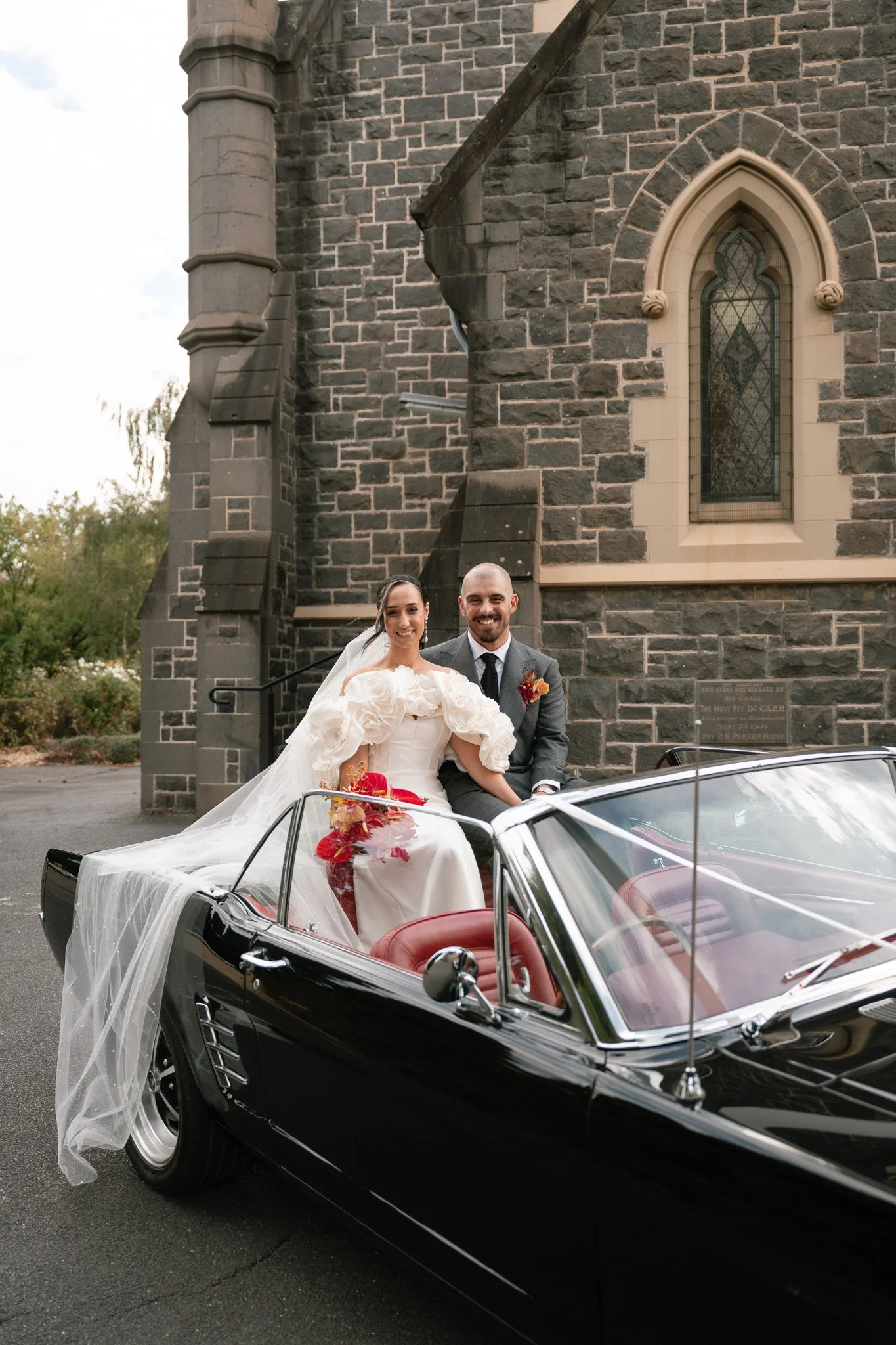 Bride and groom sitting in a black convertible car outside of a stone church, celebrating their wedding.