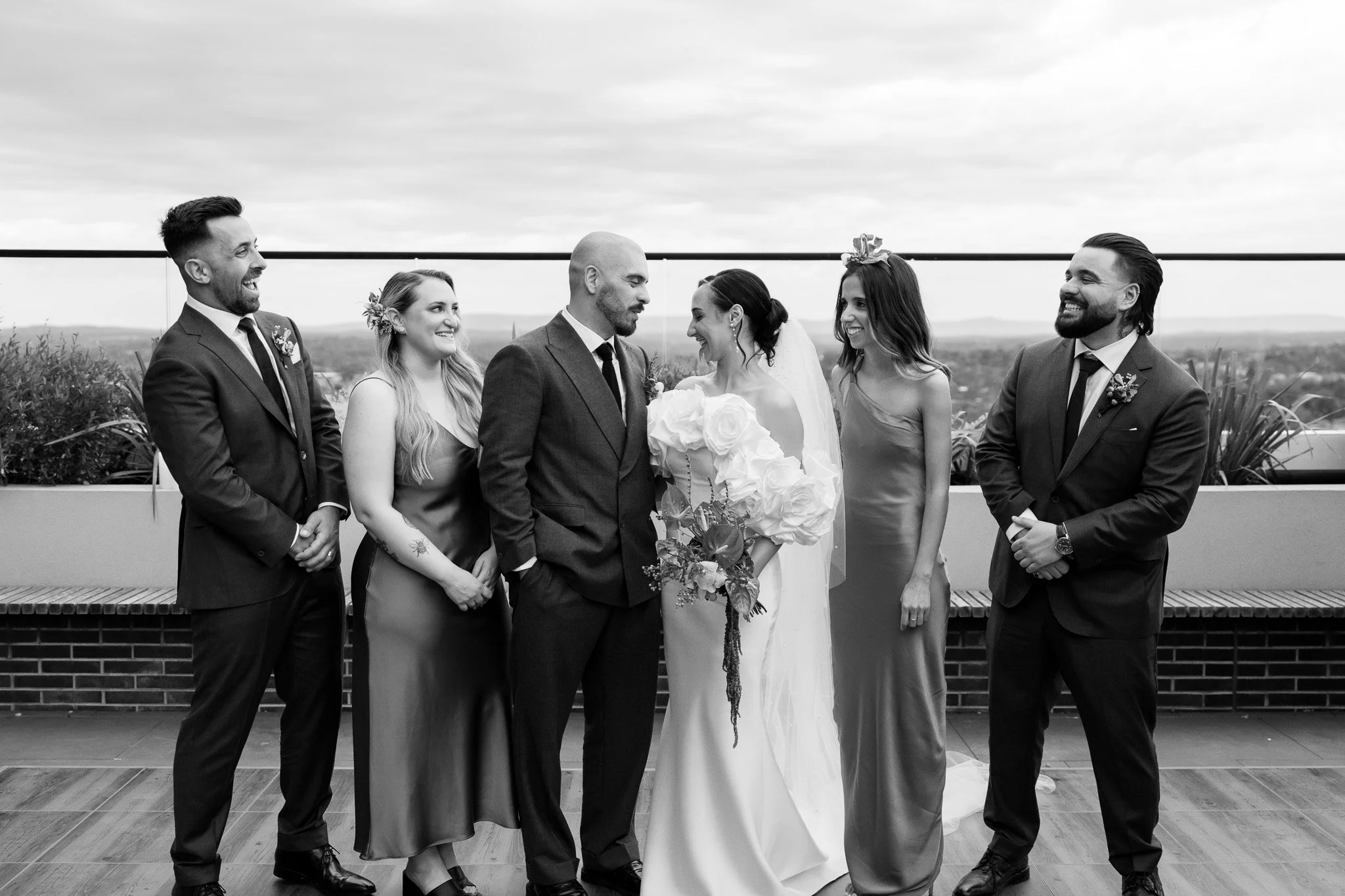 Black and white wedding photo of a bride and groom with four friends, standing outdoors on a balcony with a city view in the background.