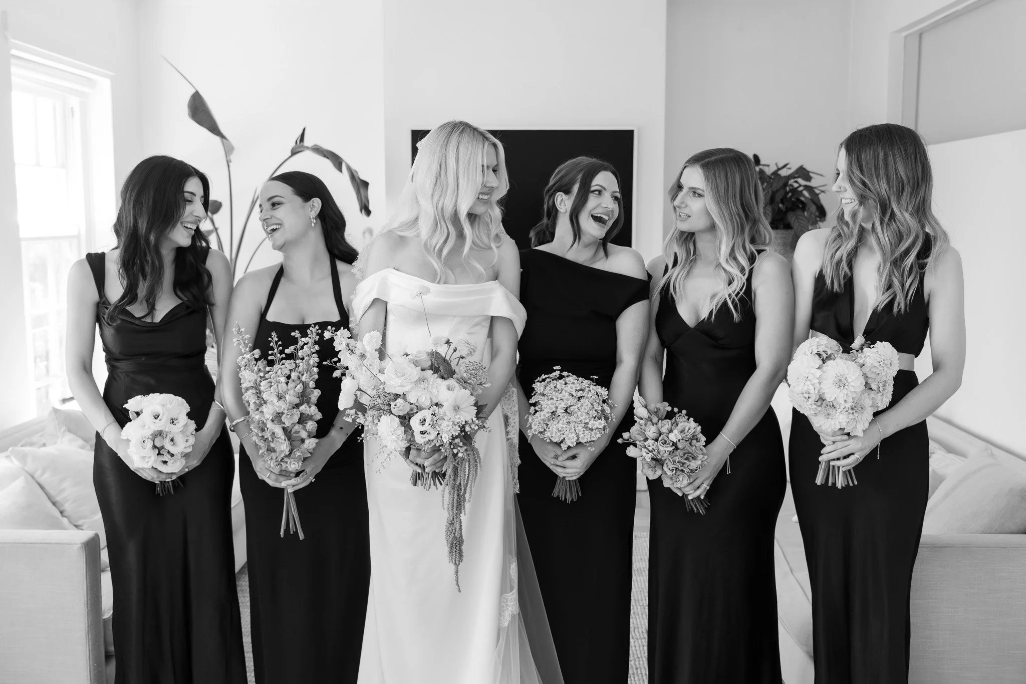 A bride in a wedding dress and her five bridesmaids in black dresses, all holding bouquets, standing in a room with a television and windows, smiling and conversing.