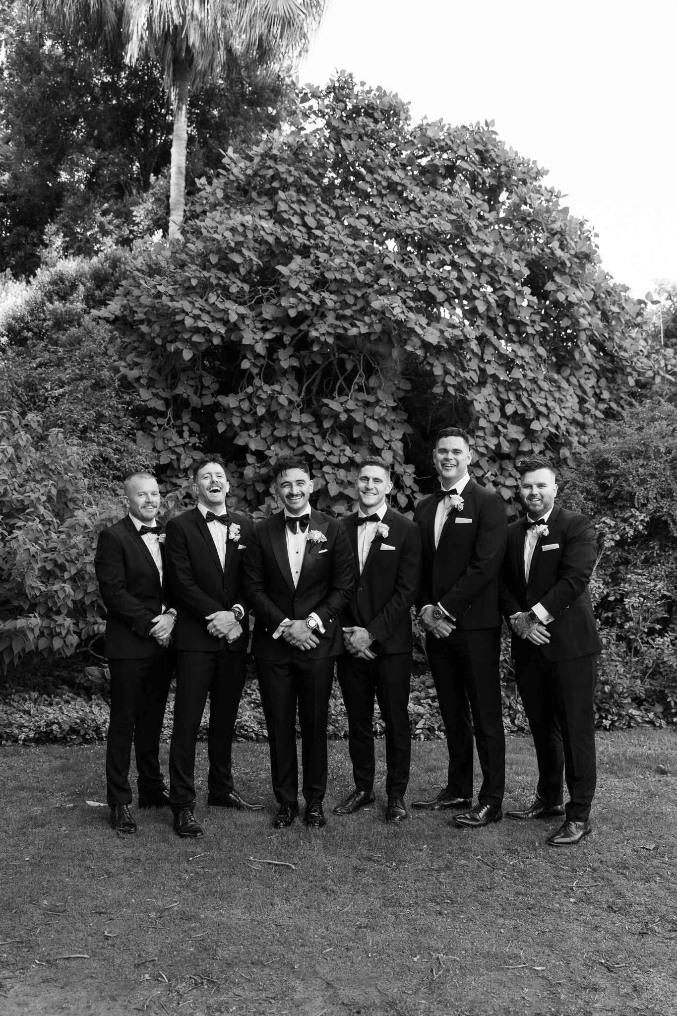 Black and white photo of seven men in tuxedos standing outdoors with greenery and a large tree behind them, smiling, likely at a formal event.
