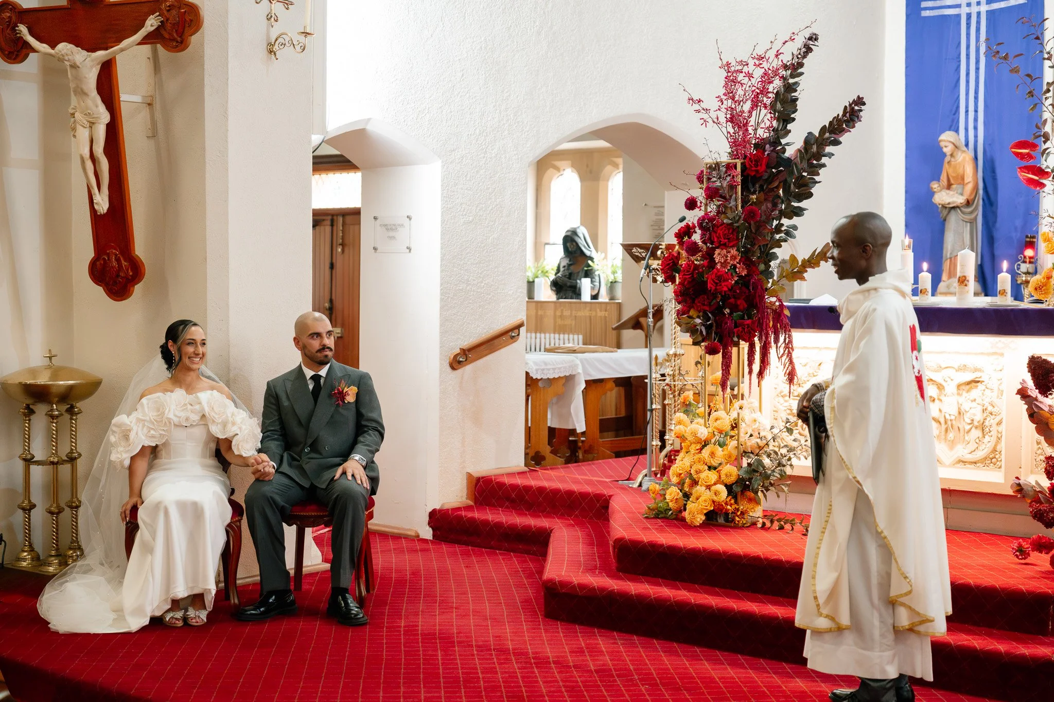 A bride and groom sitting in a church during their wedding ceremony, holding hands and smiling. The bride is in a white wedding dress with puffed sleeves, and the groom is in a dark gray suit. A priest or officiant in white robes is standing nearby. 