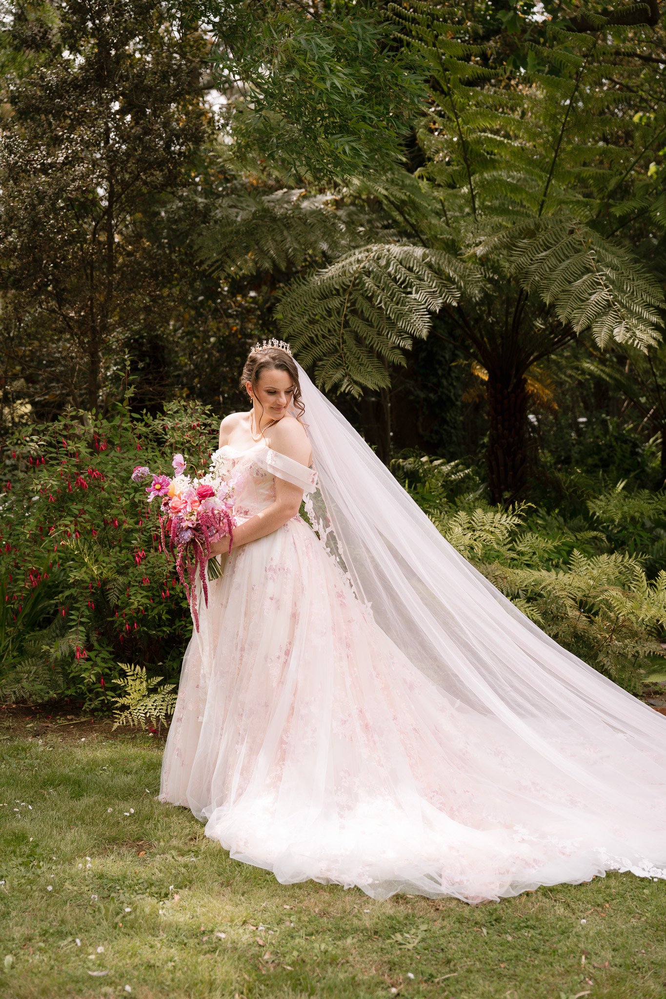 A bride in a light pink wedding gown, holding a bouquet of pink and white flowers, standing on grass in a garden surrounded by lush green plants and palm-like trees, with a long veil flowing behind her.