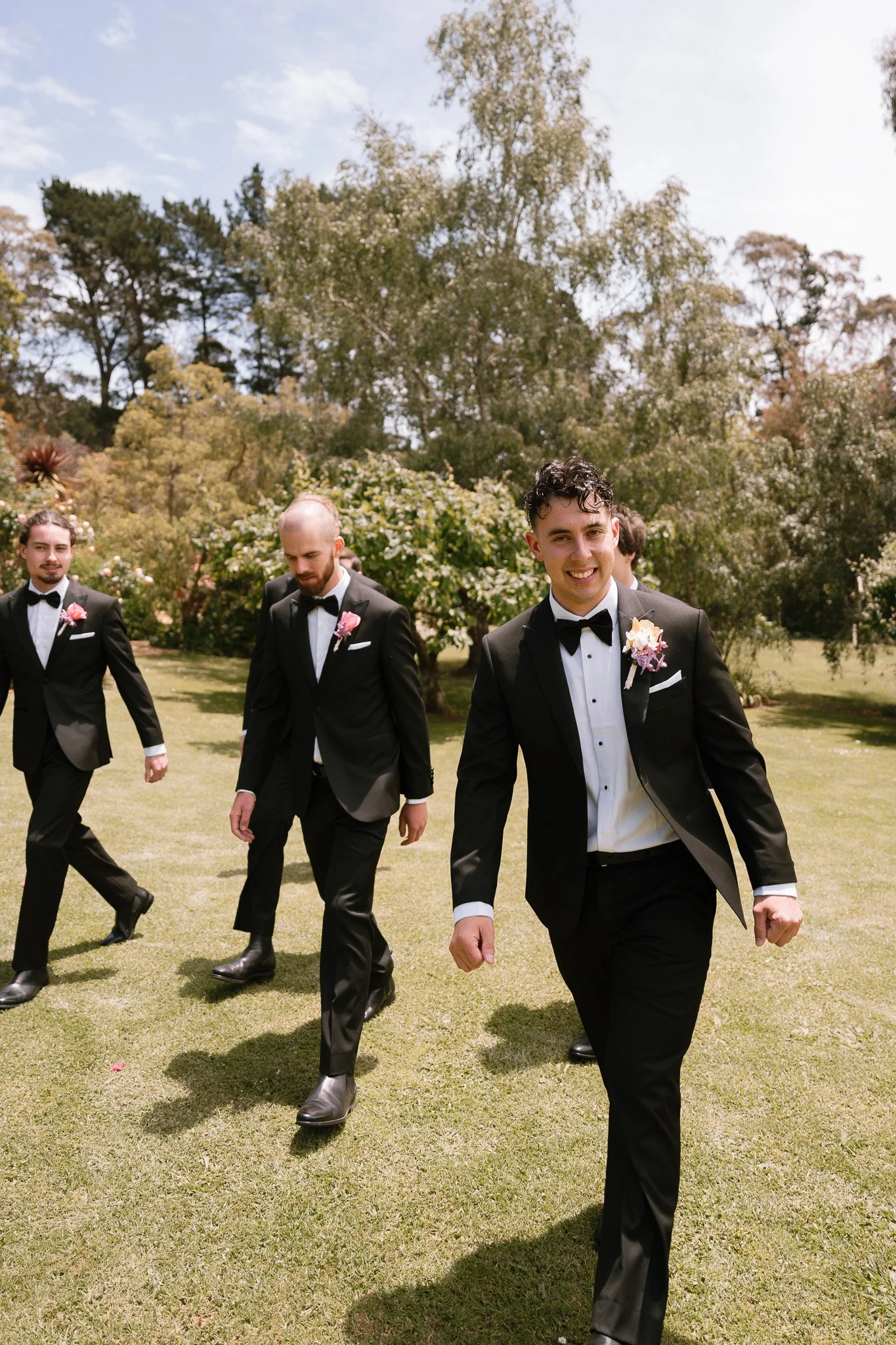 A group of four men dressed in black tuxedos with white shirts and black bow ties walking on a grassy area outdoors during the daytime. The man in front is smiling and wearing a boutonnière with purple and white flowers.