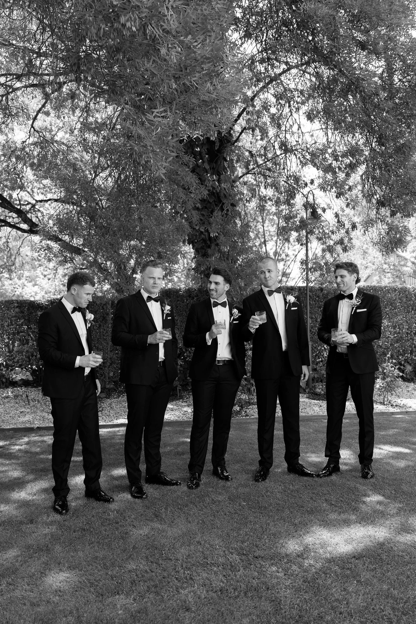 Five men in tuxedos standing outdoors on grass, holding glasses, with trees and hedges in the background, in black and white.