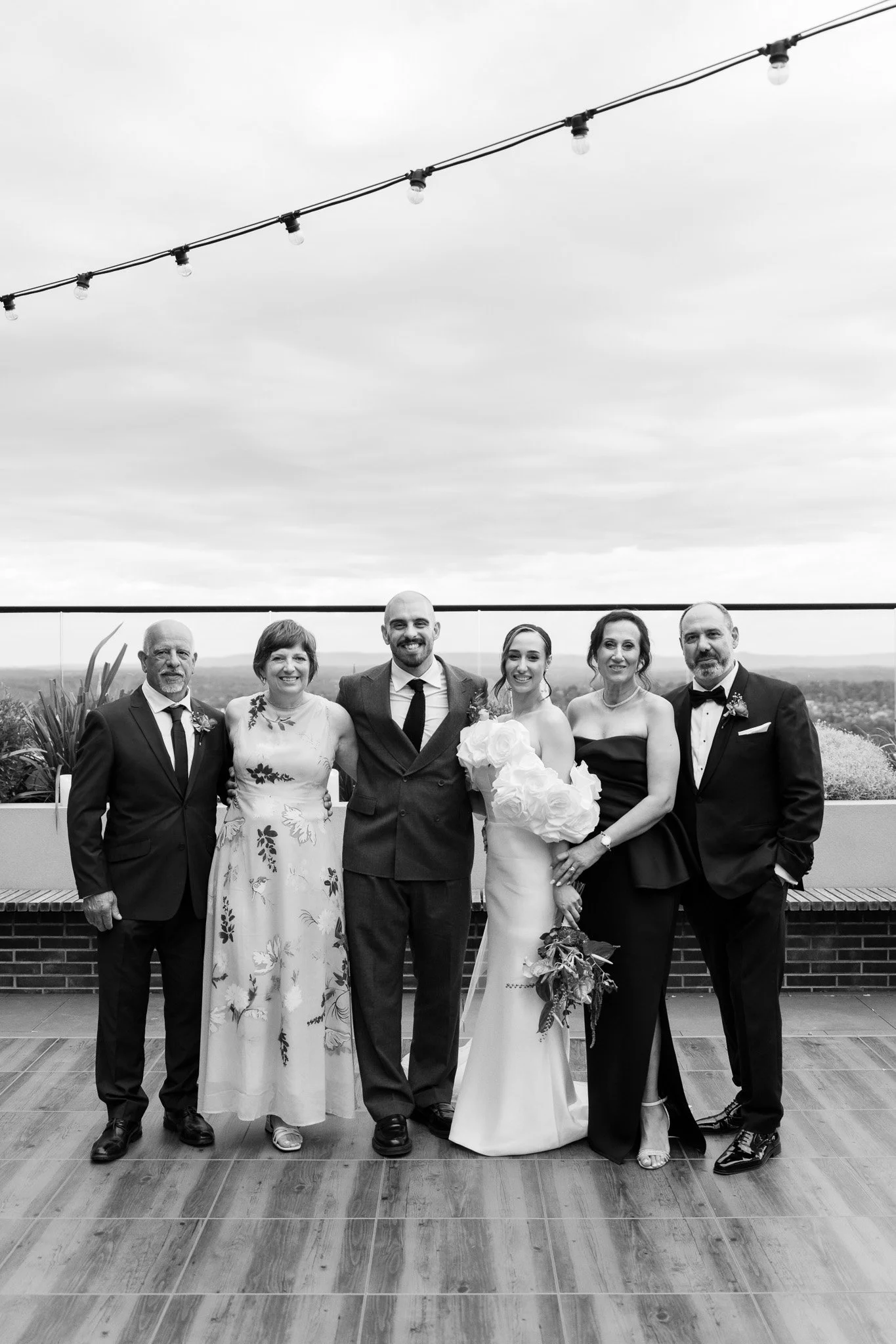 Black and white photo of a wedding celebration on a terrace with a panoramic view. A group of six people, including a bride in a white gown holding a bouquet of large flowers, and groom in a suit, are standing in the center, flanked by two men and tw