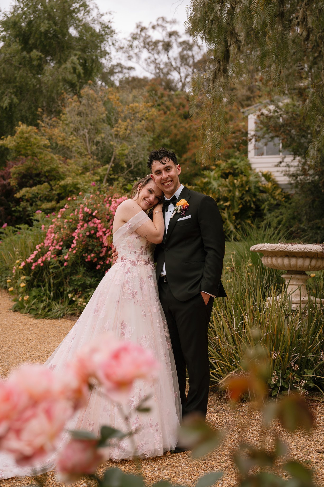 A newlywed couple embraced and smiling in a garden, with colorful flowers, greenery, and a stone birdbath in the background.