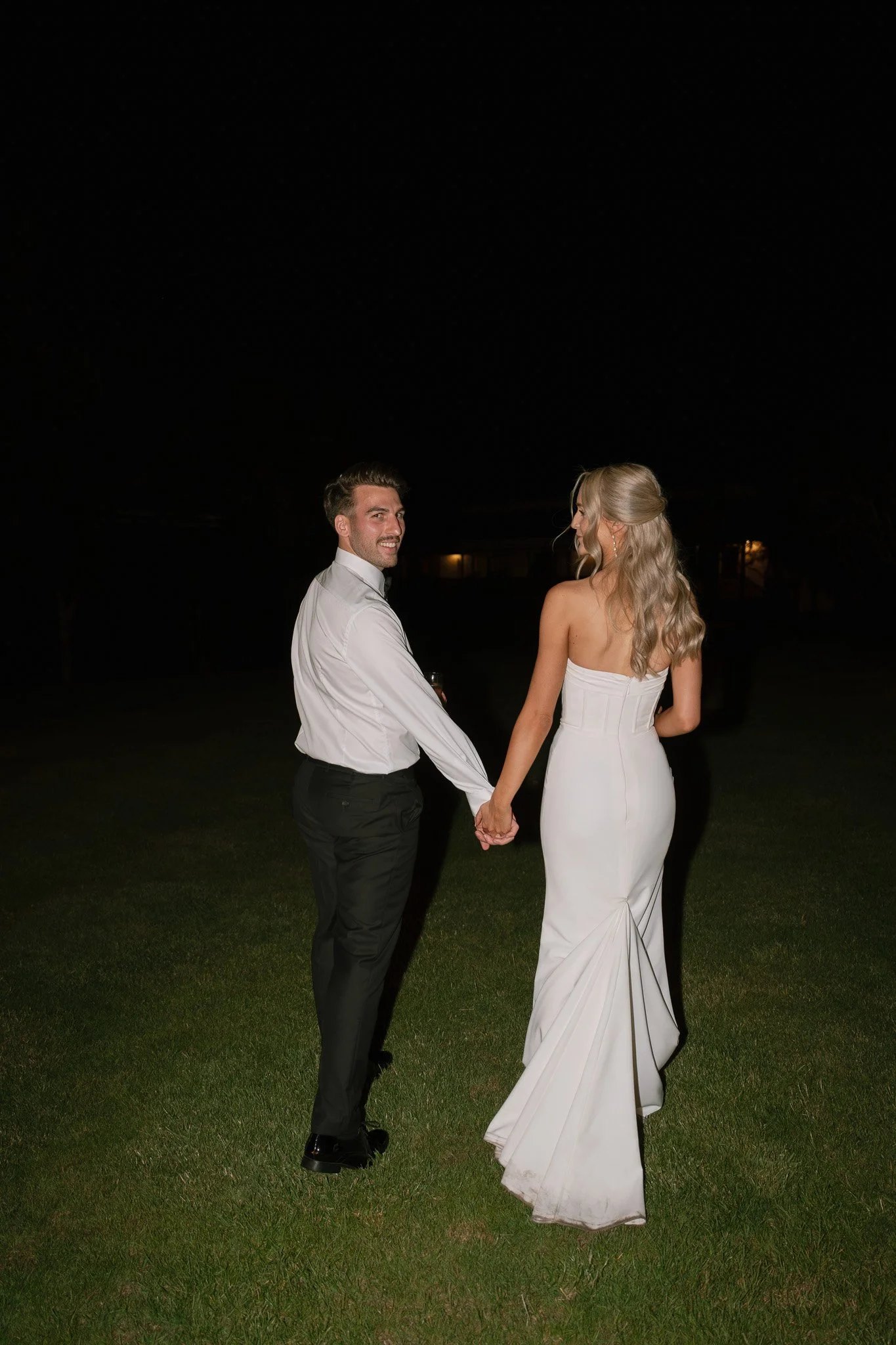 A couple dressed in formal wedding attire holding hands outdoors at night, with the man smiling and looking at the camera, and the woman turned away, wearing a strapless white gown and the man in a white shirt and black pants.
