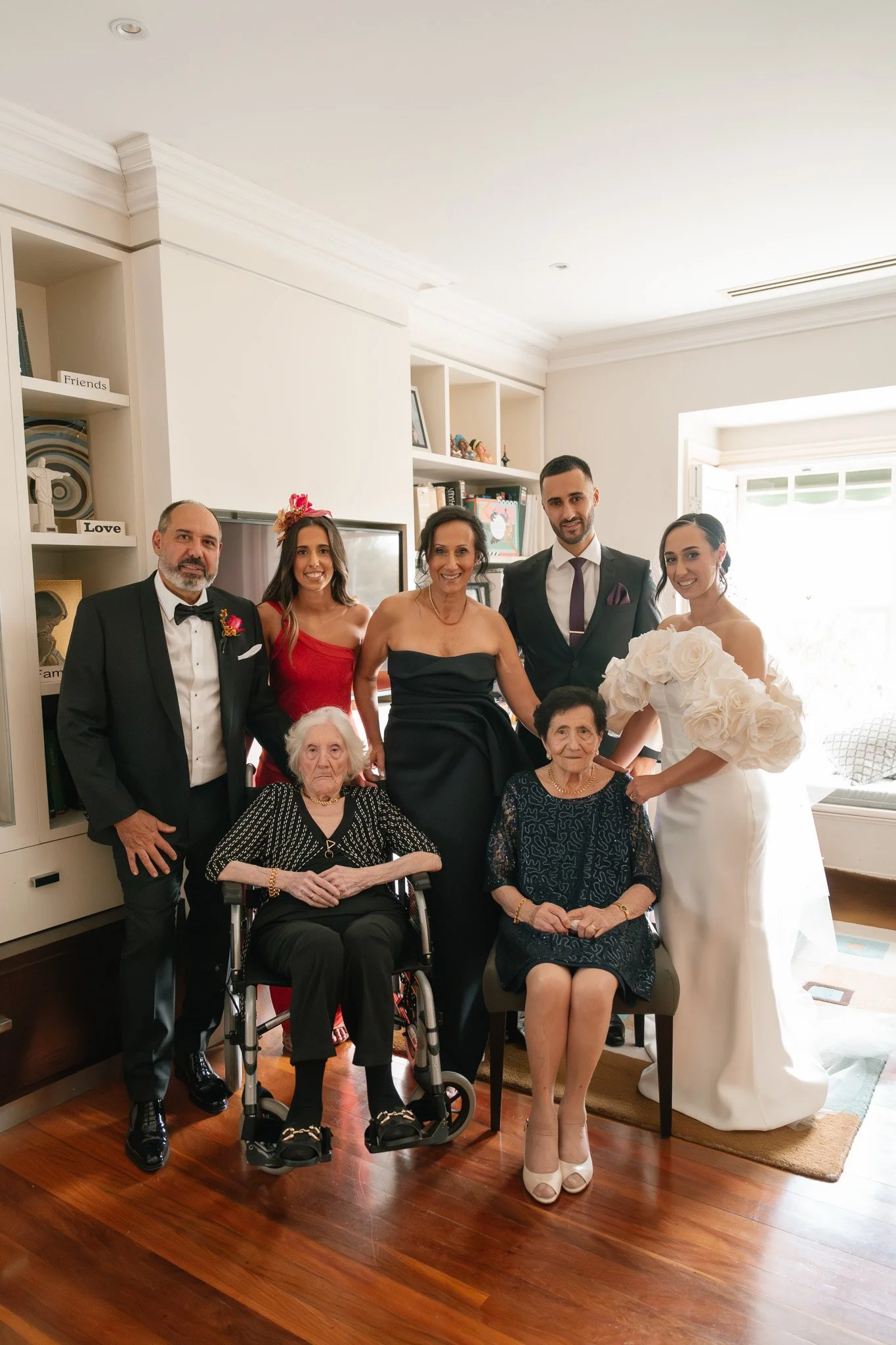 Group of people celebrating a wedding in a living room, including two elderly women seated in front, with five others standing behind them, dressed in formal attire.