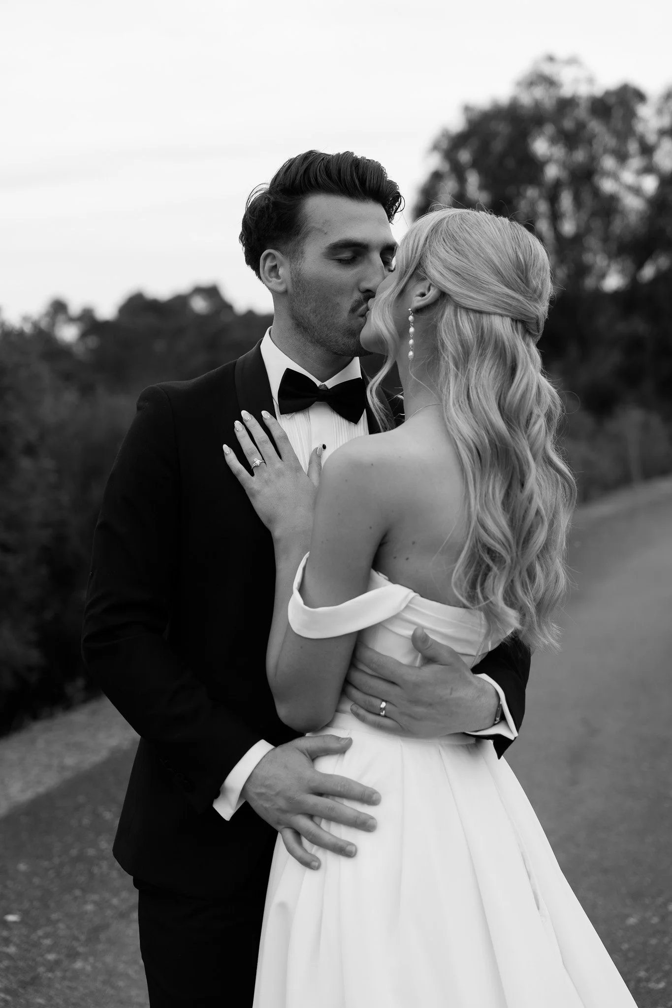 A black and white photo of a wedding couple sharing a kiss outdoors, with the groom in a tuxedo and the bride in a wedding dress.