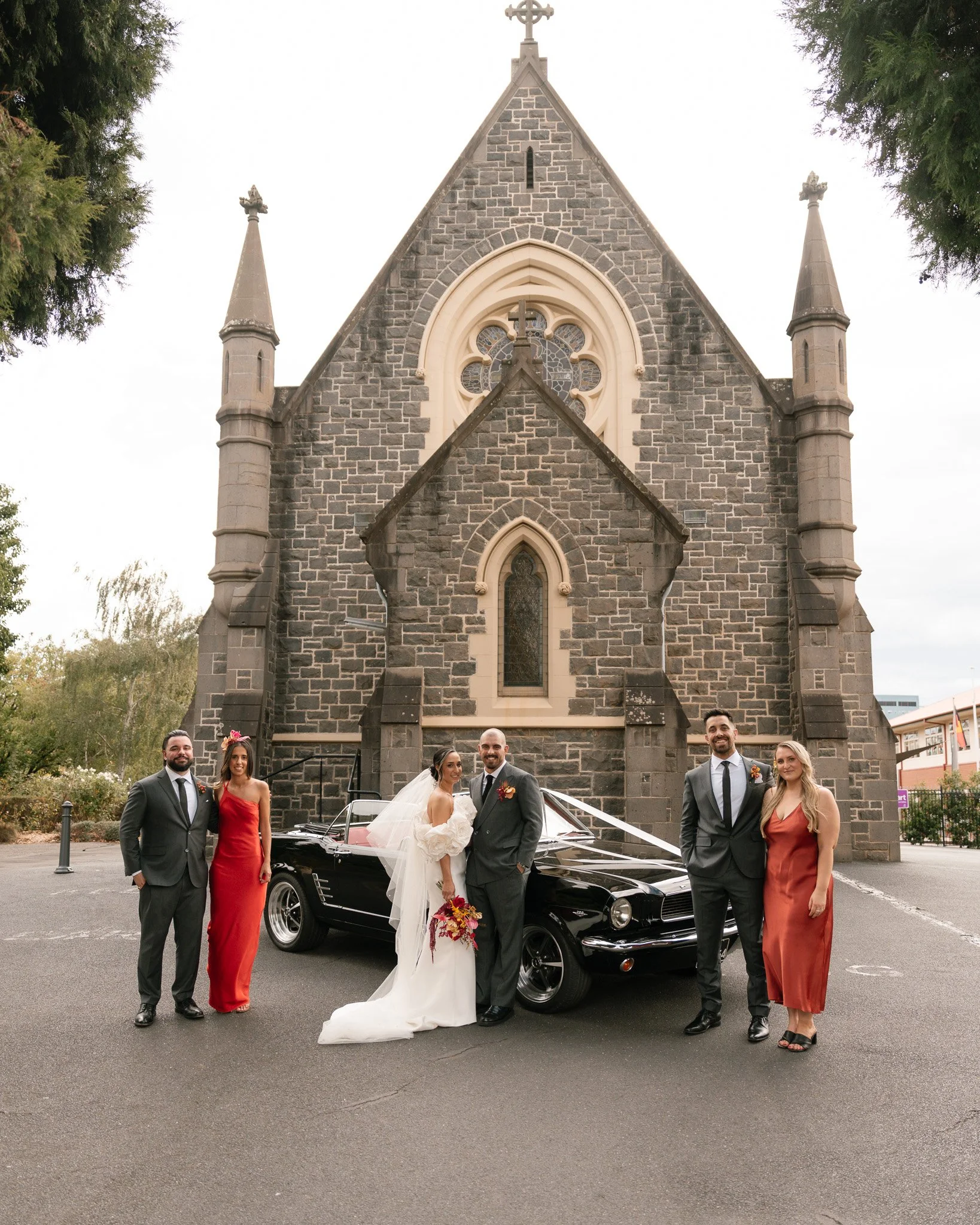 A wedding party standing in front of a black vintage car outside a stone church. The bride and groom are in the center, with the bride wearing a white wedding gown and veil, holding a bouquet, and the groom in a dark suit. Two women in red dresses an