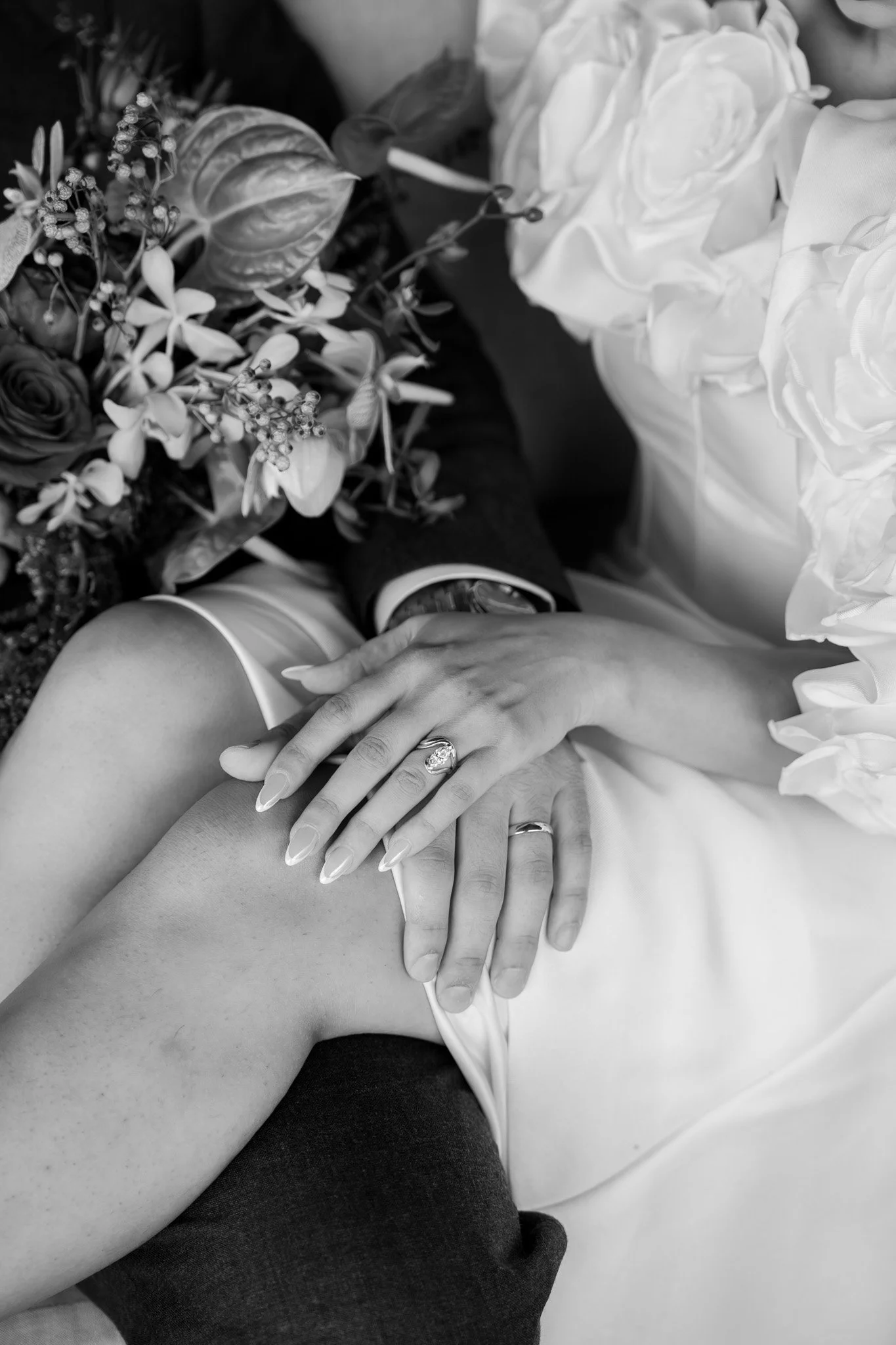 Close-up of a newlywed couple's hands resting on each other's leg, showing wedding rings. The bride's hand is on top, with a sparkling ring, and her nails are manicured. They are surrounded by wedding flowers, including roses and greenery, and dresse