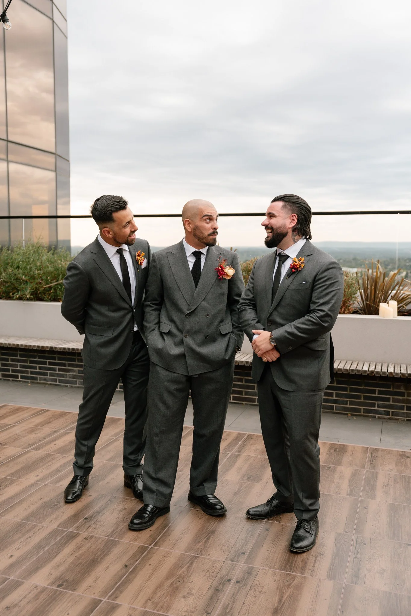 Three men dressed in suits standing on a rooftop terrace, engaged in conversation, with a city view in the background and candles on the ledge.
