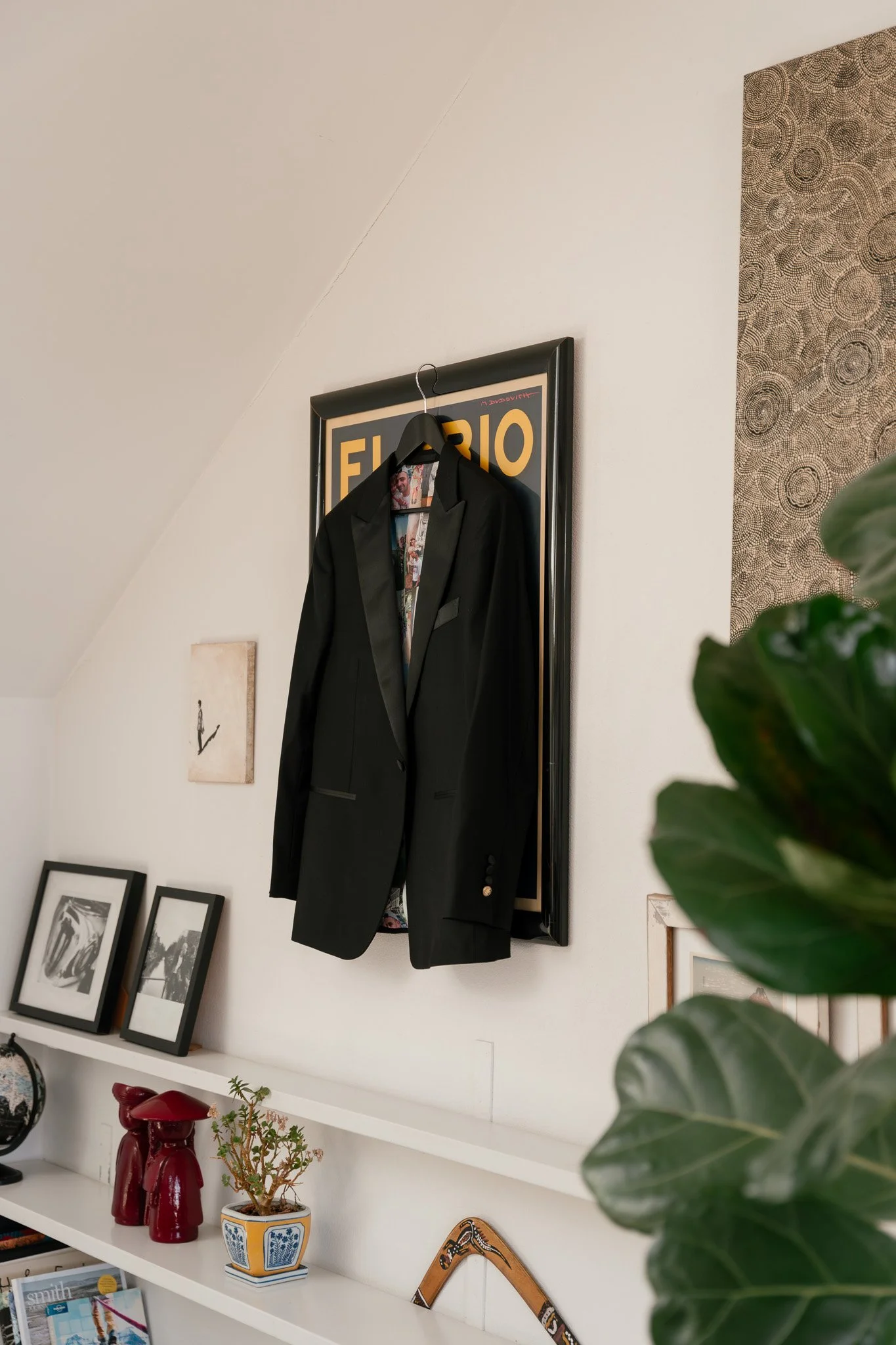 A black blazer hangs on a hanger attached to a picture frame on a white wall in a room with decorative art and plants.