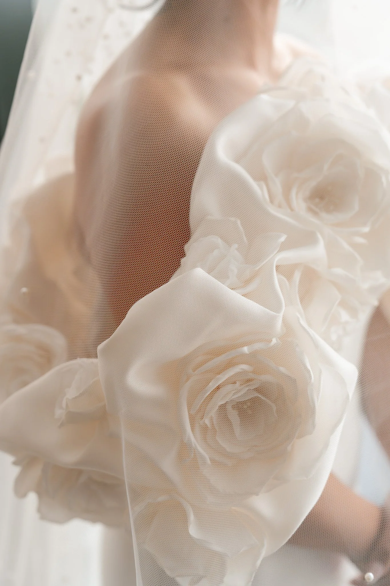 Close-up of a person behind a sheer fabric embroidered with large, white roses, with only part of their shoulder and neck visible.