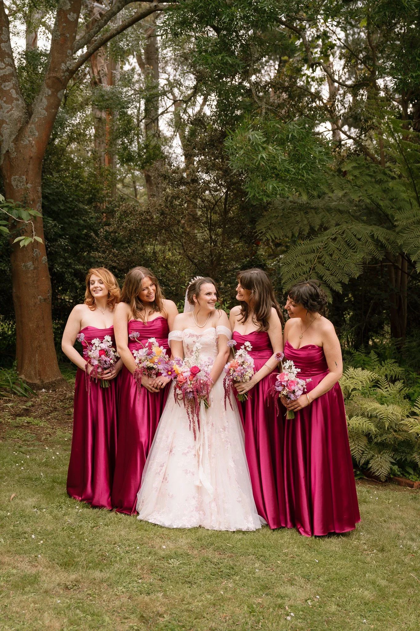 A bride in a white wedding dress with pink floral accents standing with five bridesmaids in magenta strapless gowns, all holding bouquets of pink and purple flowers, outdoors in a lush, green wooded area.