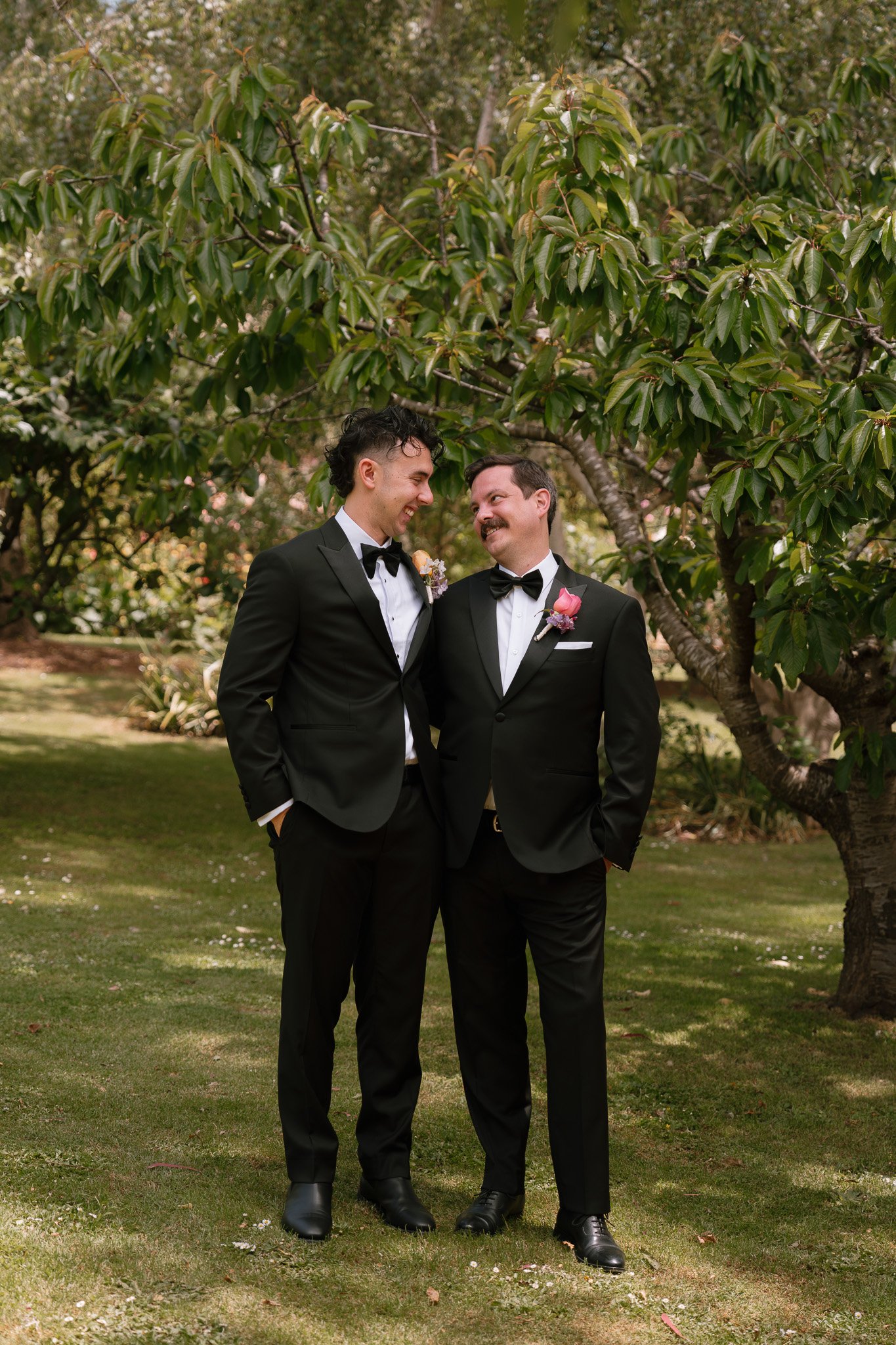 Two men in tuxedos smiling at each other outdoors under a tree.