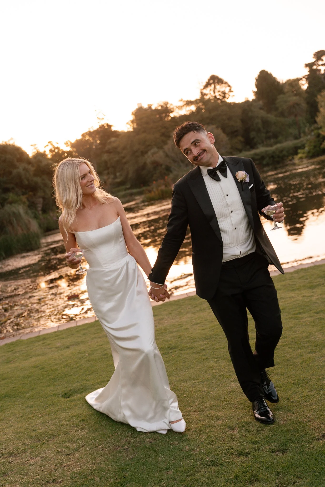 A bride and groom holding hands and walking on grass near a lake during sunset with trees in the background. The bride is wearing a white strapless wedding gown and the groom is in a black tuxedo with a bow tie, holding a glass of champagne.