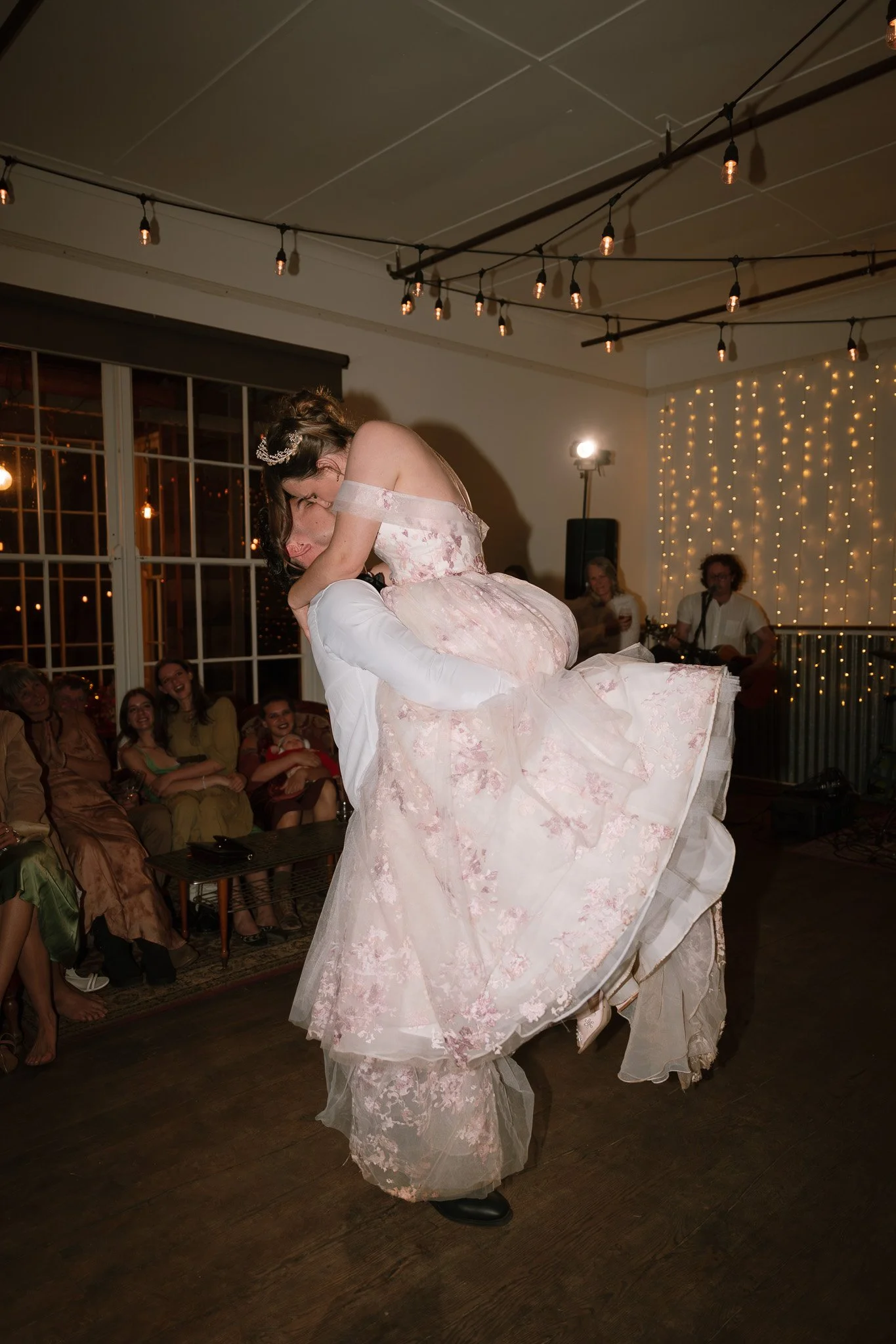 A bride being lifted and spun by a groom during a wedding reception, with friends watching and smiling in the background, decorated with string lights.