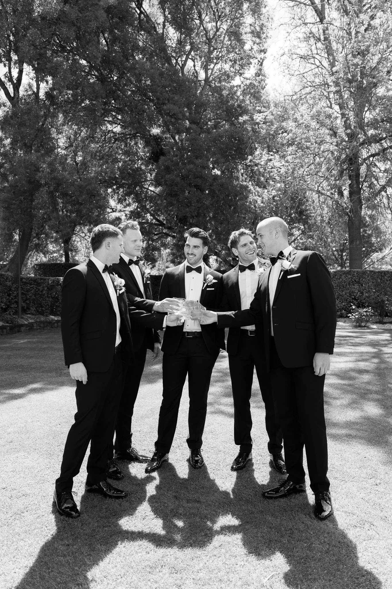 Six men dressed in tuxedos holding a trophy together outdoors, smiling, with trees in the background.