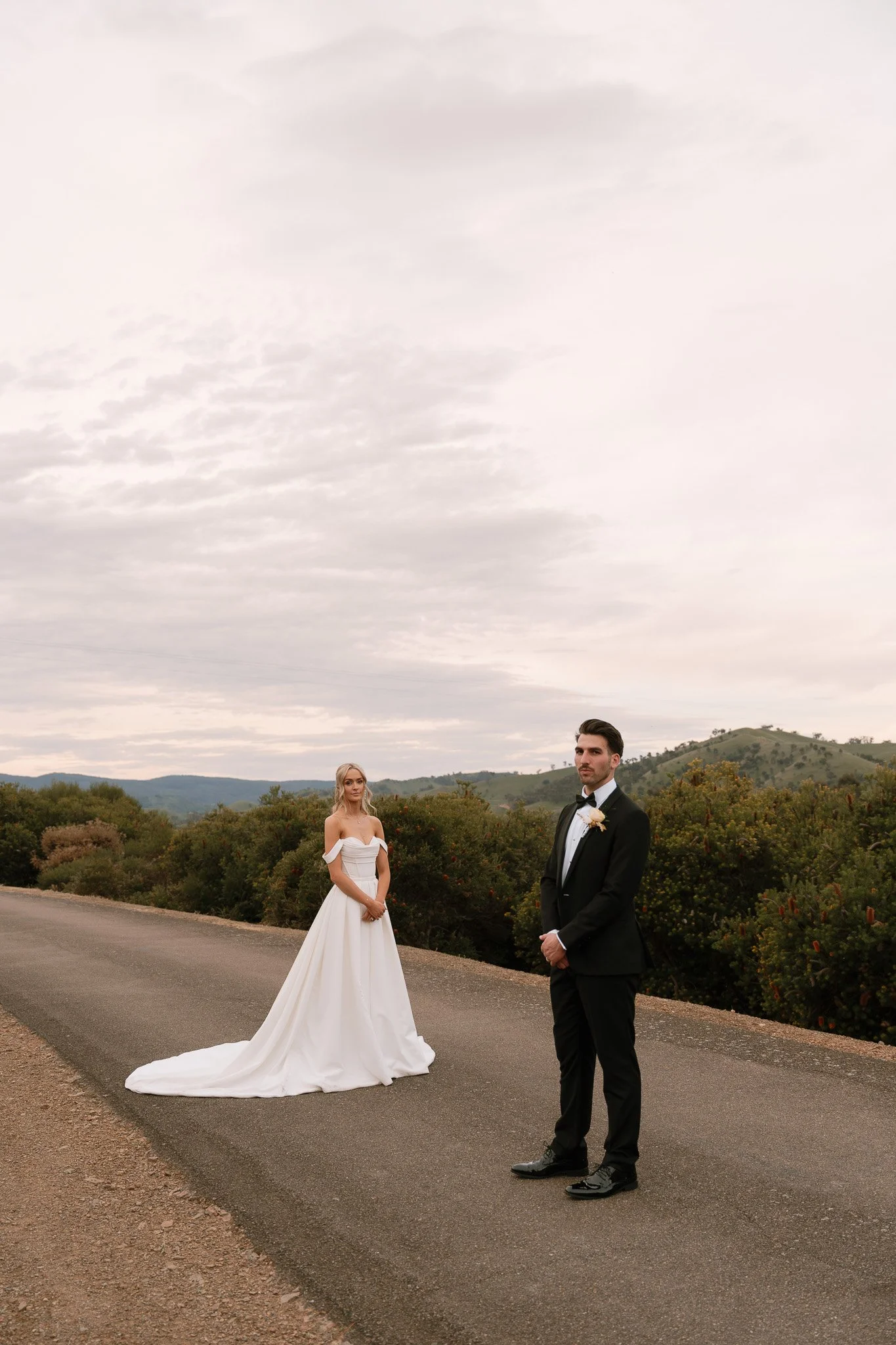A bride and groom standing on a rural road with a scenic mountainous landscape in the background during a wedding photoshoot.