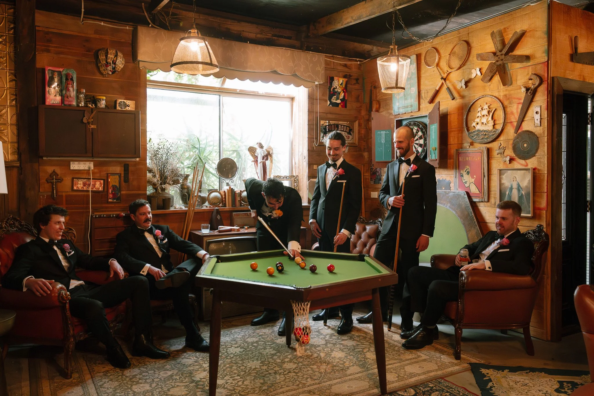 Six men in tuxedos playing and socializing around a pool table in a wood-paneled room with vintage decor.