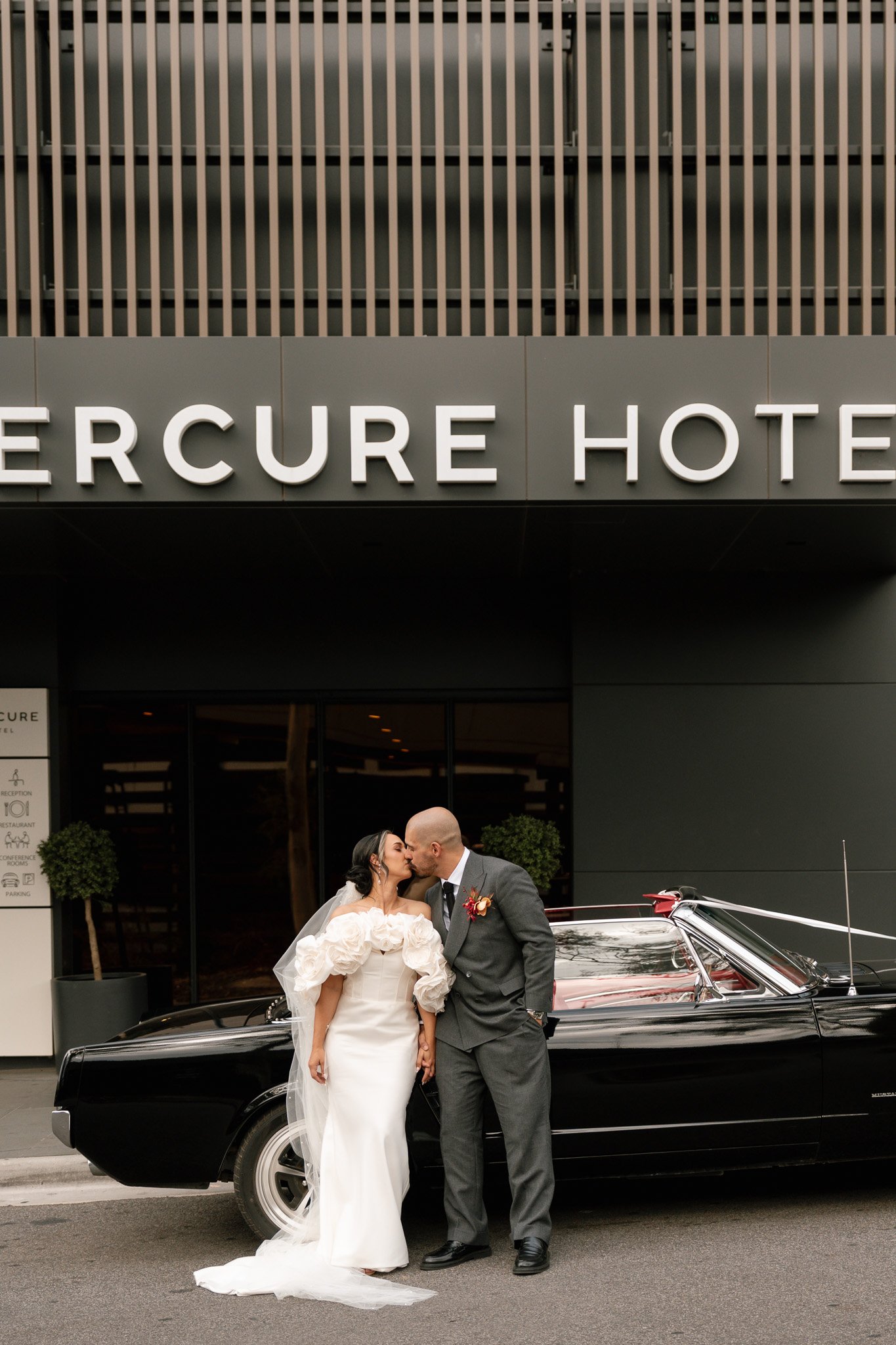 A newlywed couple sharing a kiss in front of a black vintage car outside the Mercure Hotel, with the hotel's sign visible above them.