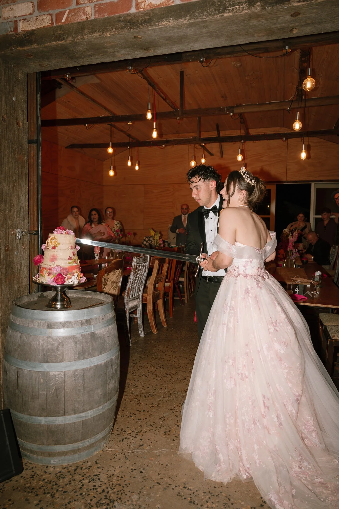 A bride and groom cutting a wedding cake together in a rustic venue with exposed brick and wooden ceiling, surrounded by wedding guests.