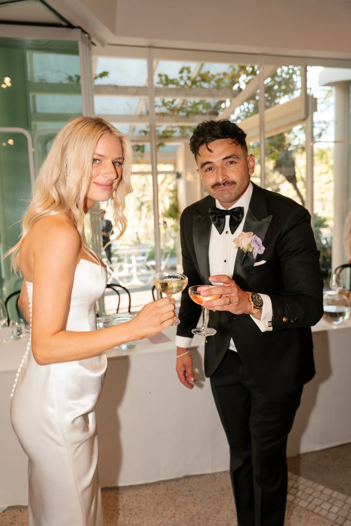 A man in a tuxedo with a bow tie and a woman in a white satin dress holding champagne glasses at a celebration.