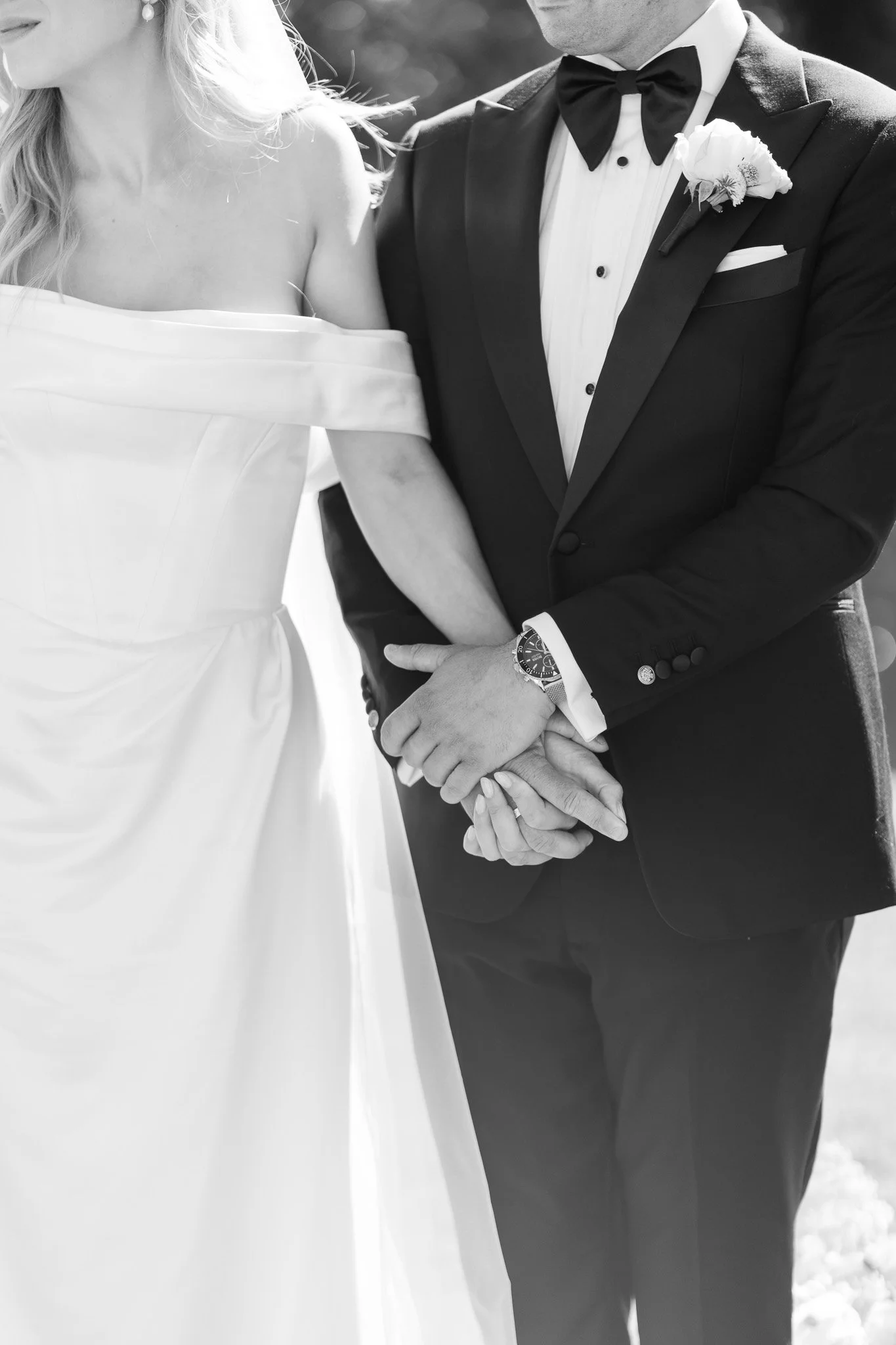 Black and white photo of a bride and groom holding hands during their wedding, with the bride in a strapless dress and the groom in a tuxedo with a bow tie and boutonniere.