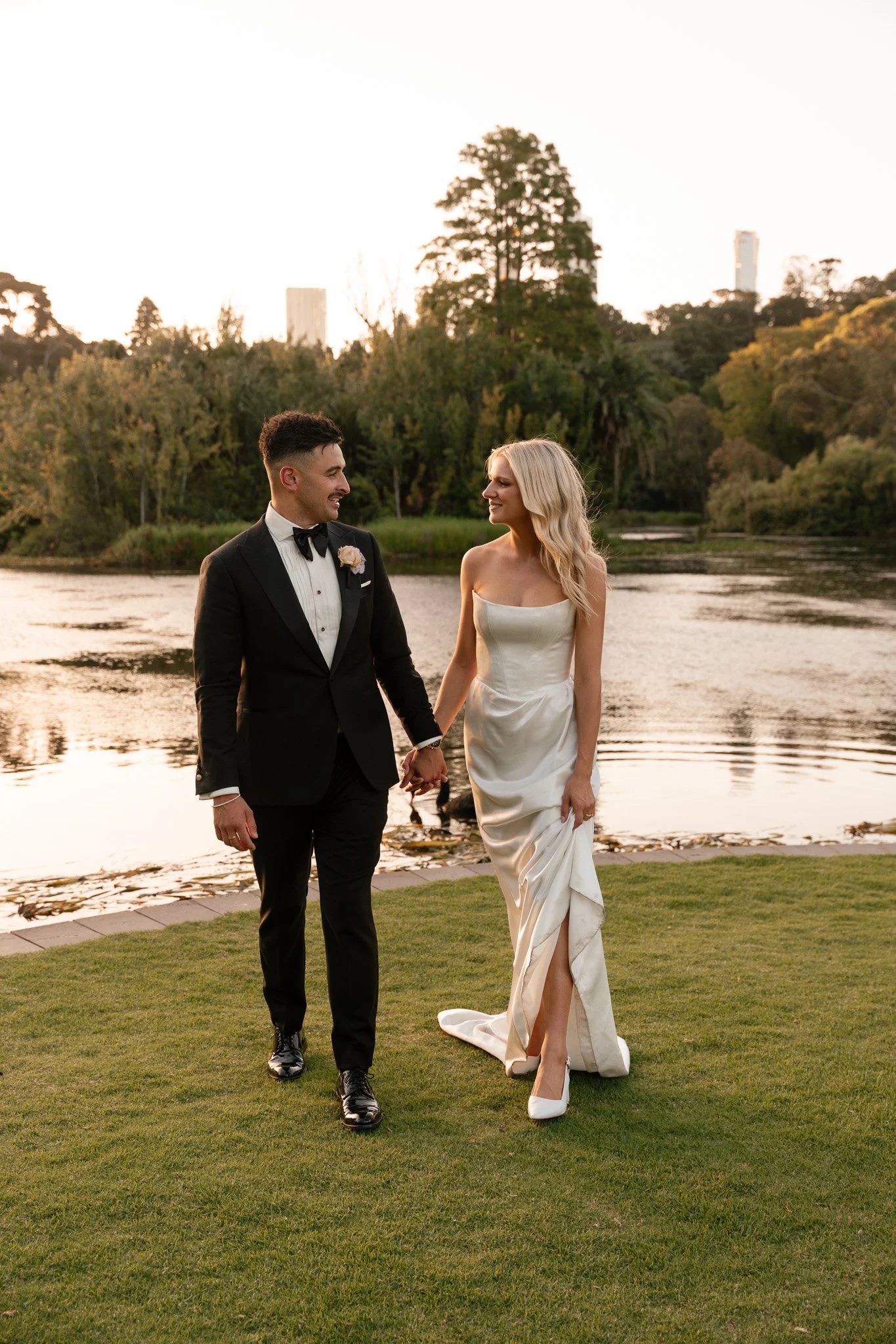 A wedding couple walking hand in hand by a river during sunset.