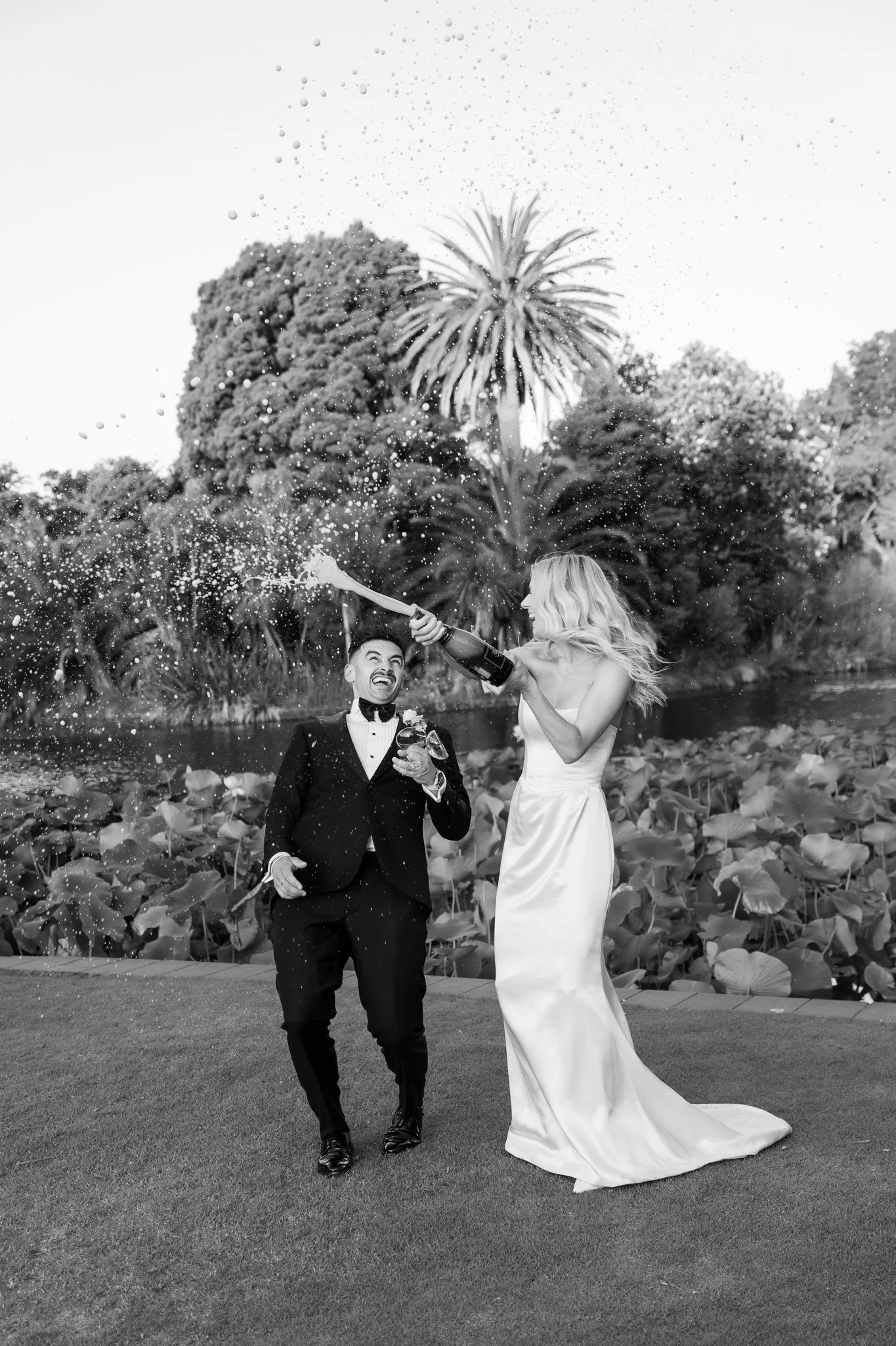 A black and white photo of a newlywed couple celebrating by a pond, with the bride spraying champagne on the groom.