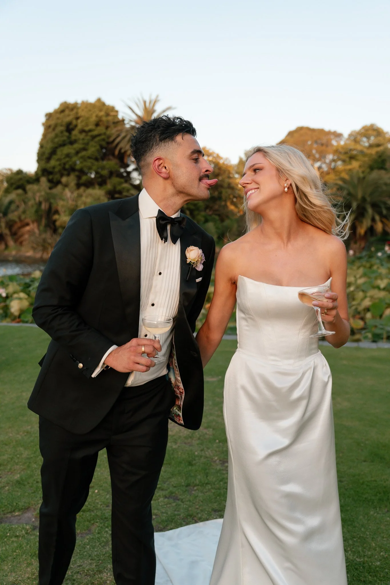 A newlywed couple at an outdoor wedding reception, dressed in wedding attire, holding glasses of champagne, standing on a lawn with trees and a lake in the background.