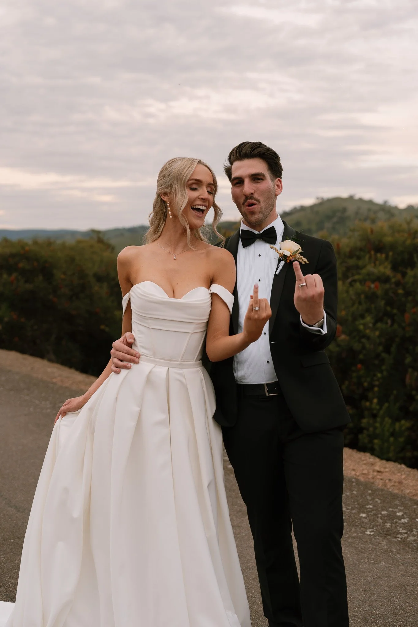A bride and groom in wedding attire standing outdoors, both showing middle fingers at the camera.