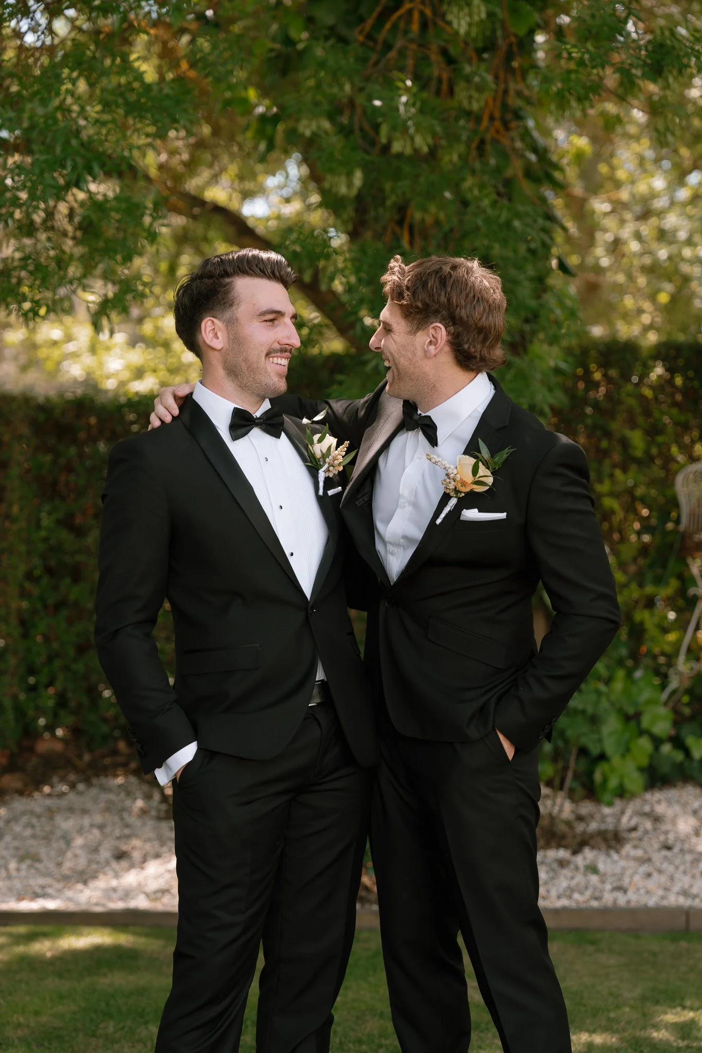 Two men in tuxedos smiling and looking at each other, outside in a garden with trees and greenery.