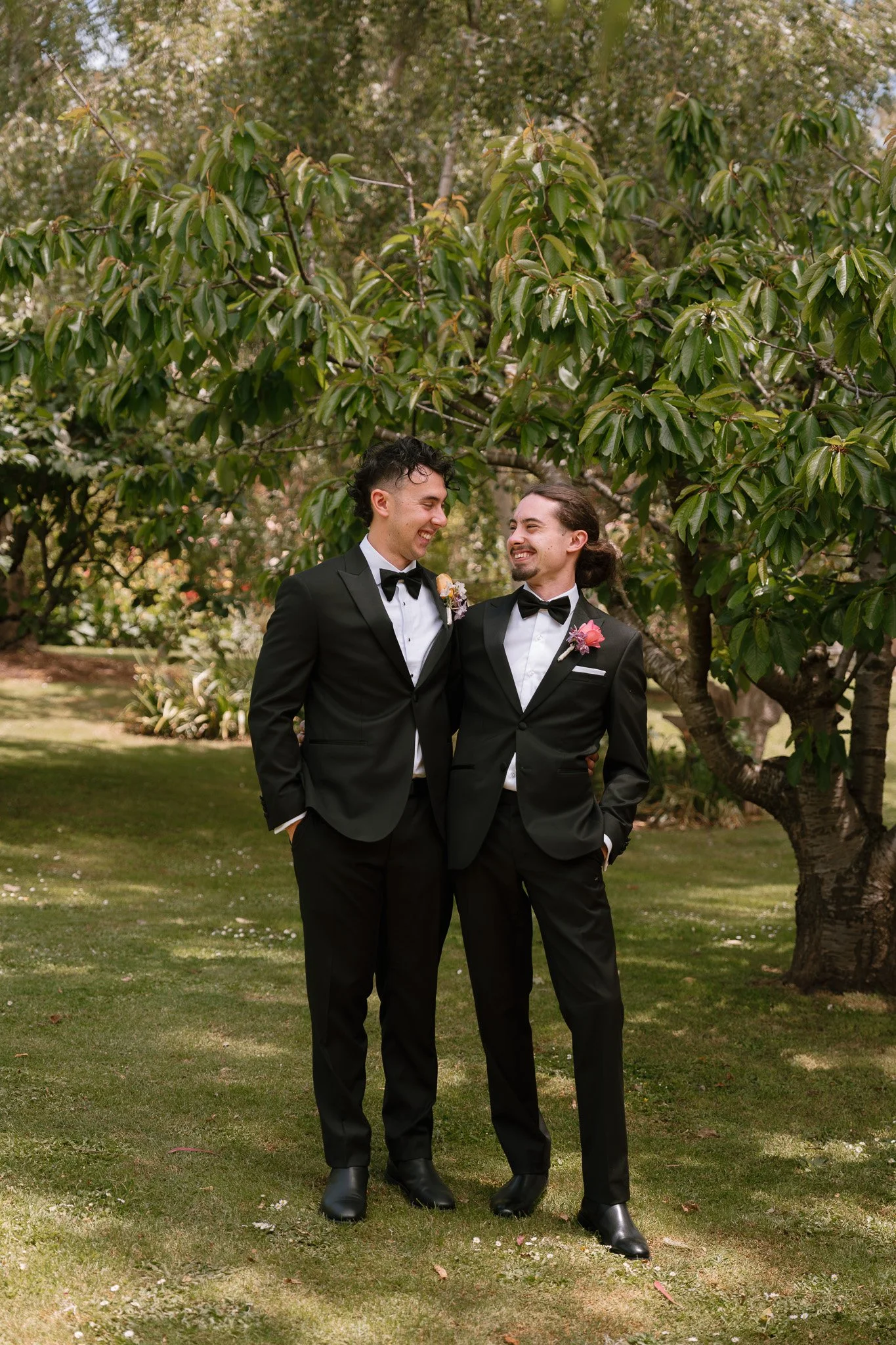 Two men in tuxedos standing on grass in a garden, smiling and looking at each other, with a large green tree in the background.