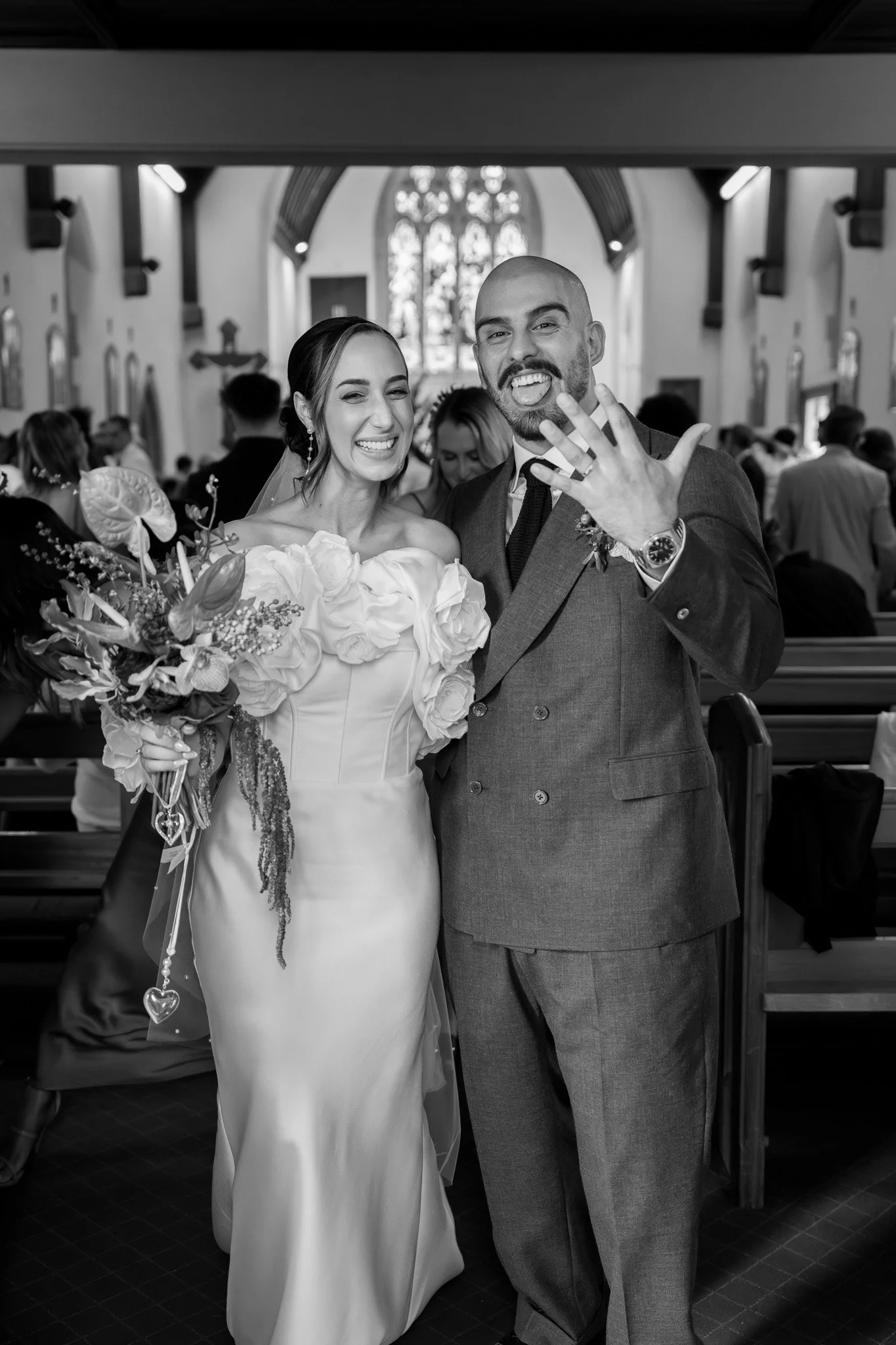 Black and white photo of a bride and groom inside a church, smiling and showing off wedding rings, surrounded by guests.