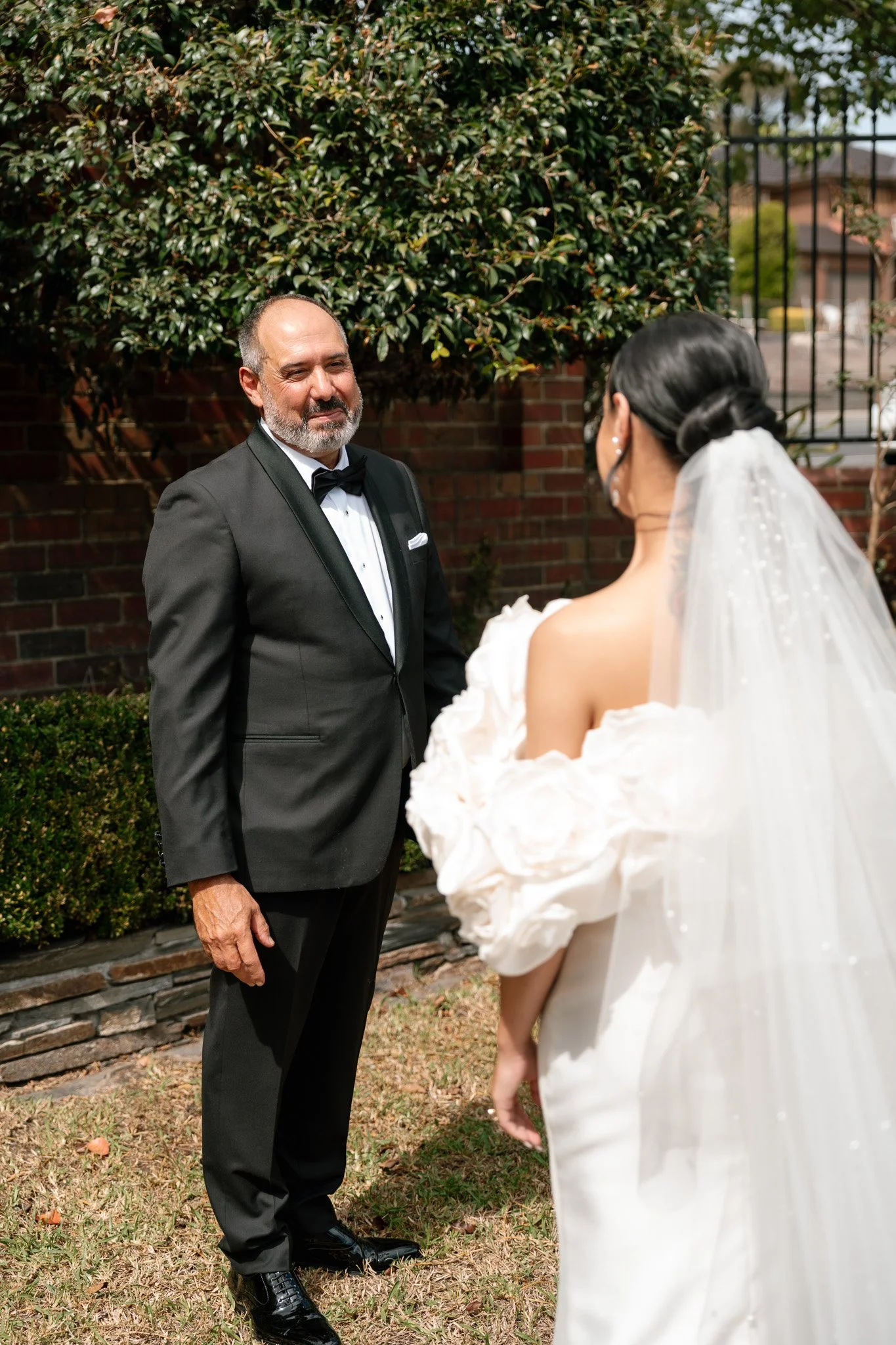 An older man in a tuxedo is smiling and looking at a woman in a white wedding dress with a veil, outside in front of a brick wall and greenery.